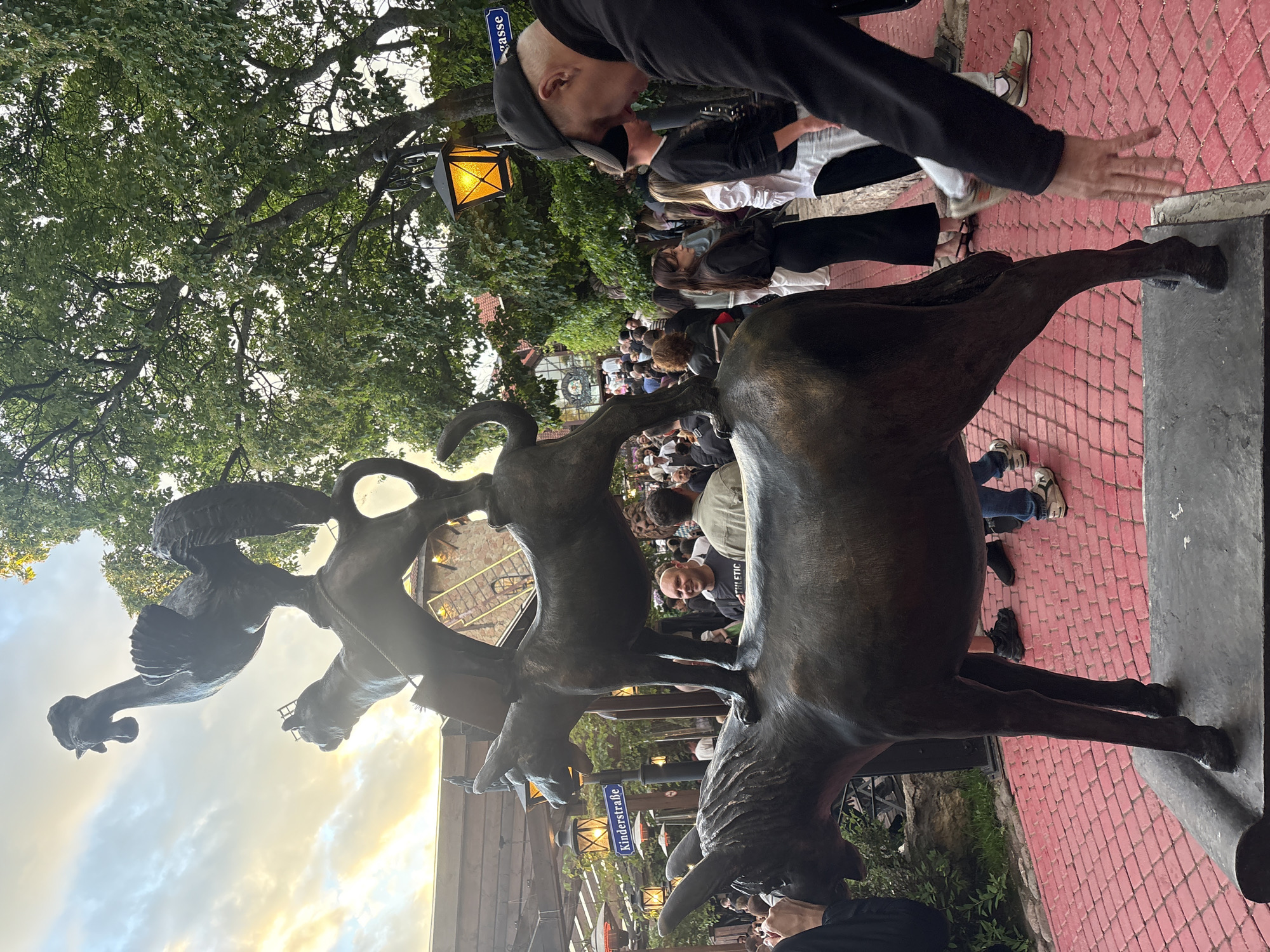 The image depicts a bronze sculpture of four animals stacked on top of each other: a donkey at the base, followed by a dog, a cat, and a rooster at the top. The sculpture is located in a public outdoor space, likely a pedestrian area, with red brick paving. In the background, there are people gathered, suggesting it is a popular spot. Trees with green foliage overhang the scene, and streetlights are visible, one of which is illuminated with a warm yellow light. A street sign with the text "Kinderstraße" and "gasse" is also present, indicating a street name in German, which is incongruous with the stated location of Saint Petersburg, Russia. A man wearing a black baseball cap and black jacket is in the foreground, his arm extended towards the sculpture. The overall atmosphere appears to be that of a lively gathering during daytime or early evening.