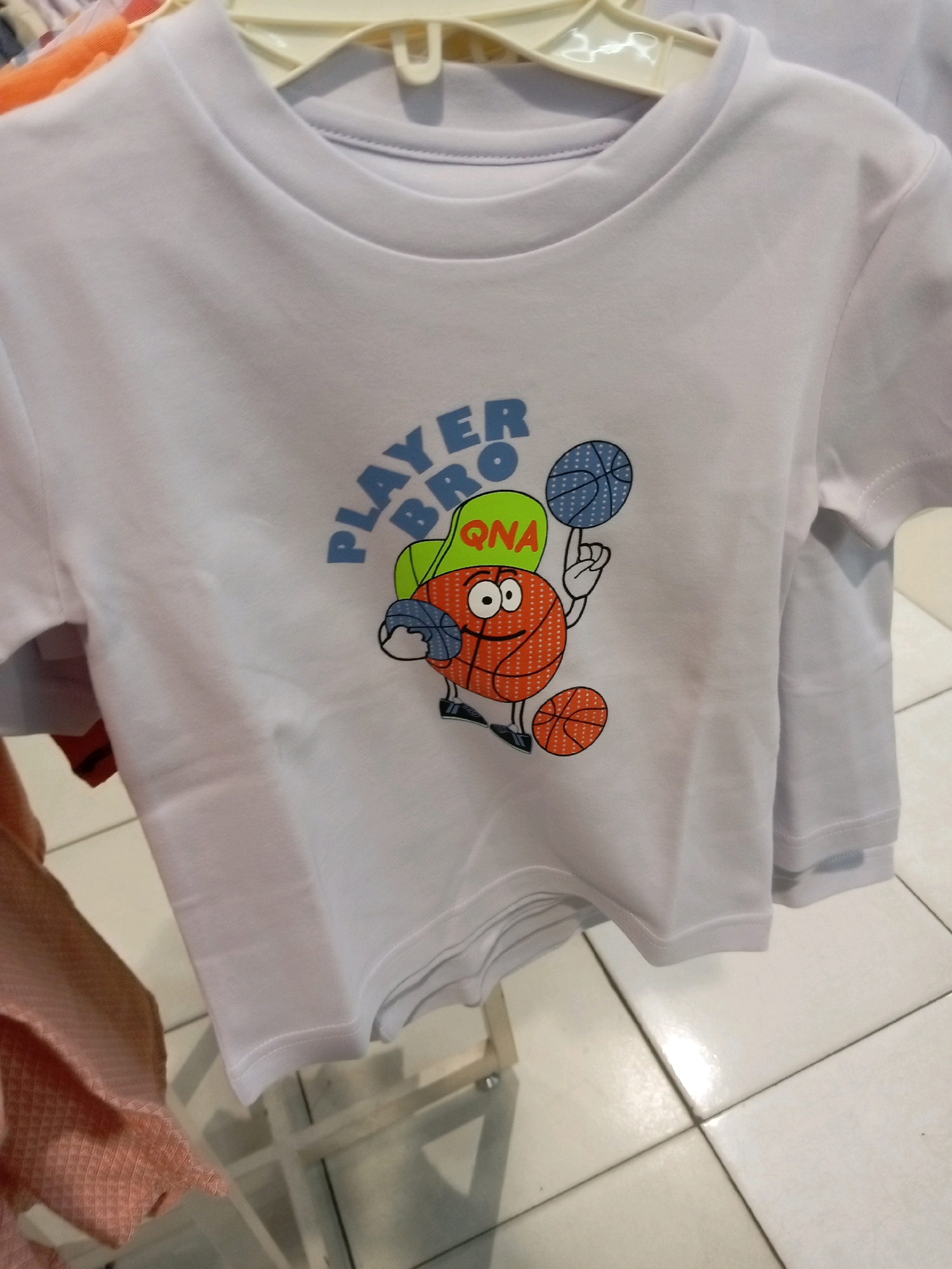 A white t-shirt with a cartoon graphic is displayed on a hanger. The graphic depicts a basketball character wearing a green hat with the text "QNA" inside it. The character is holding one basketball and has another at its feet. The text "PLAYER BRO" is printed above the character in blue letters. The t-shirt is hanging on a white hanger, and other clothing items can be seen in the background. The t-shirt is likely located in a retail store in Batam, Indonesia, given the context.