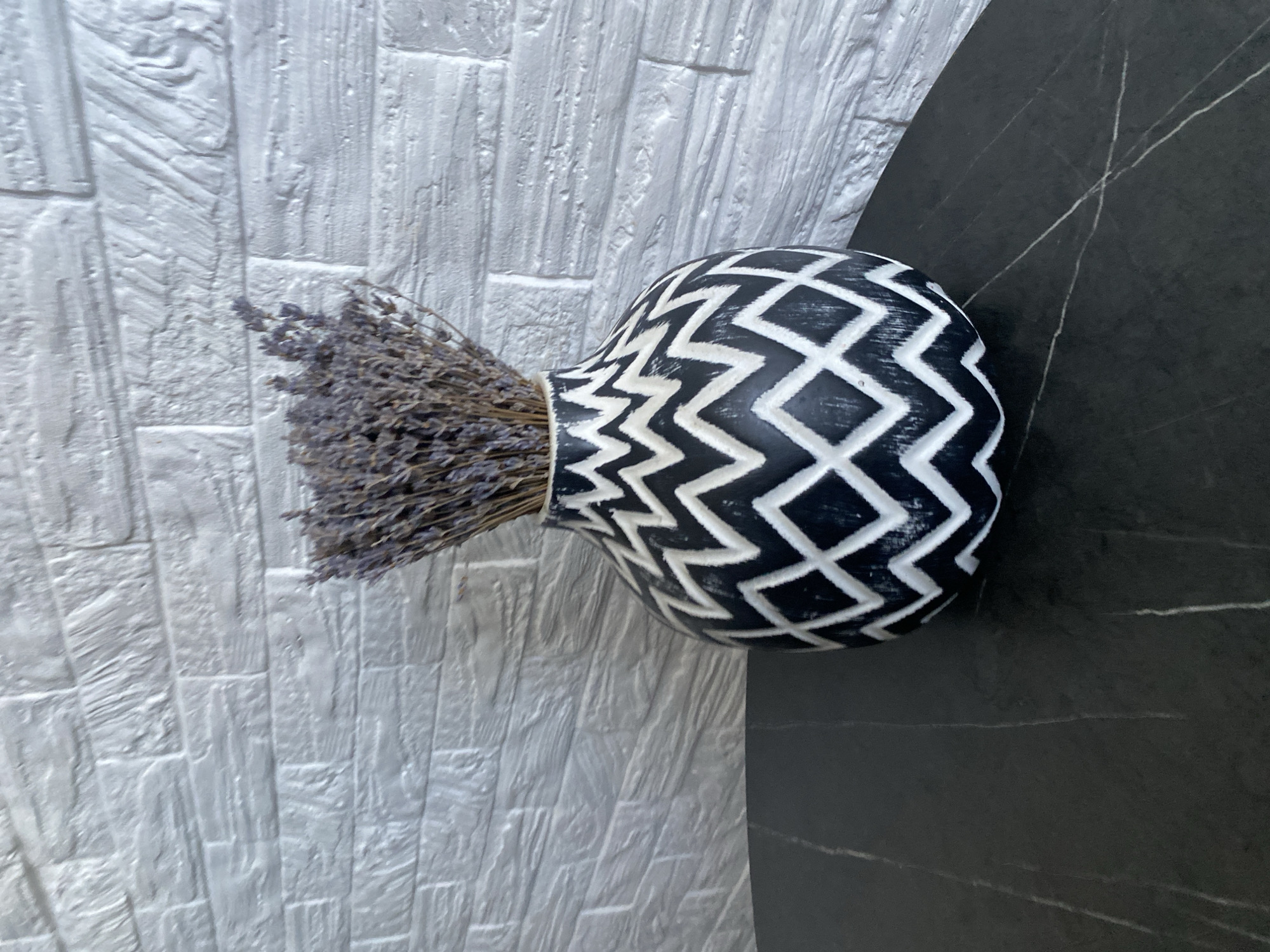 A black and white patterned vase filled with dried lavender sits on a dark marble table, set against a textured white brick wall. The vase has a bold, zig-zag pattern in black and white, and the lavender is a muted purple. The table surface is reflective, with subtle lighter veining visible. The brick wall behind the vase has a rough, uneven texture. The image is a still life composition, likely indoors. No people or actions are depicted. The location in Chișinău, Moldova is not specified.
