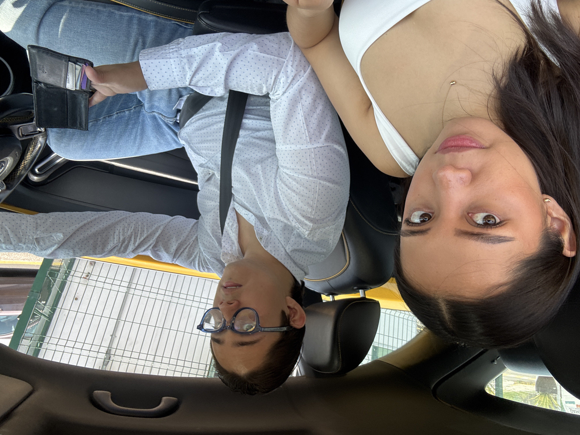 Inside a vehicle, a man and a woman are seen oriented at a 180-degree angle to the camera. The man is on the left wearing glasses, a button-down shirt, jeans, and a seatbelt. He holds a black wallet with credit cards visible. The woman, positioned on the right, wears a white top and has long dark hair. The background shows a glimpse of an exterior scene through the car window: a metal fence and some greenery, implying an outdoor environment, presumably the city of Puebla, Mexico.