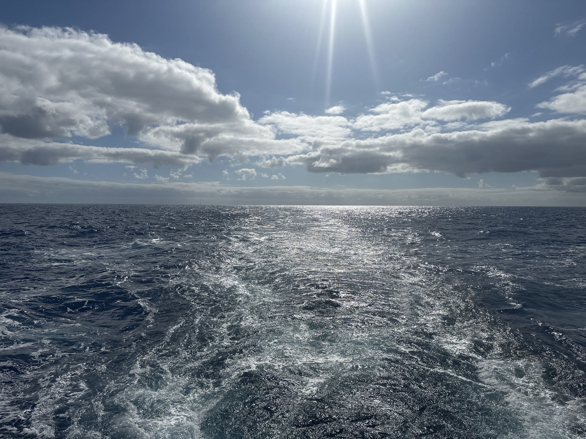 The image depicts an open ocean scene under a partly cloudy sky with the sun shining brightly. The water is a deep blue, with white foamy waves and the wake of a boat visible in the foreground. The sky is a light blue, with puffy white clouds scattered throughout. The sun is near the top of the frame, casting a bright glare and beams of light. The location is the open ocean.