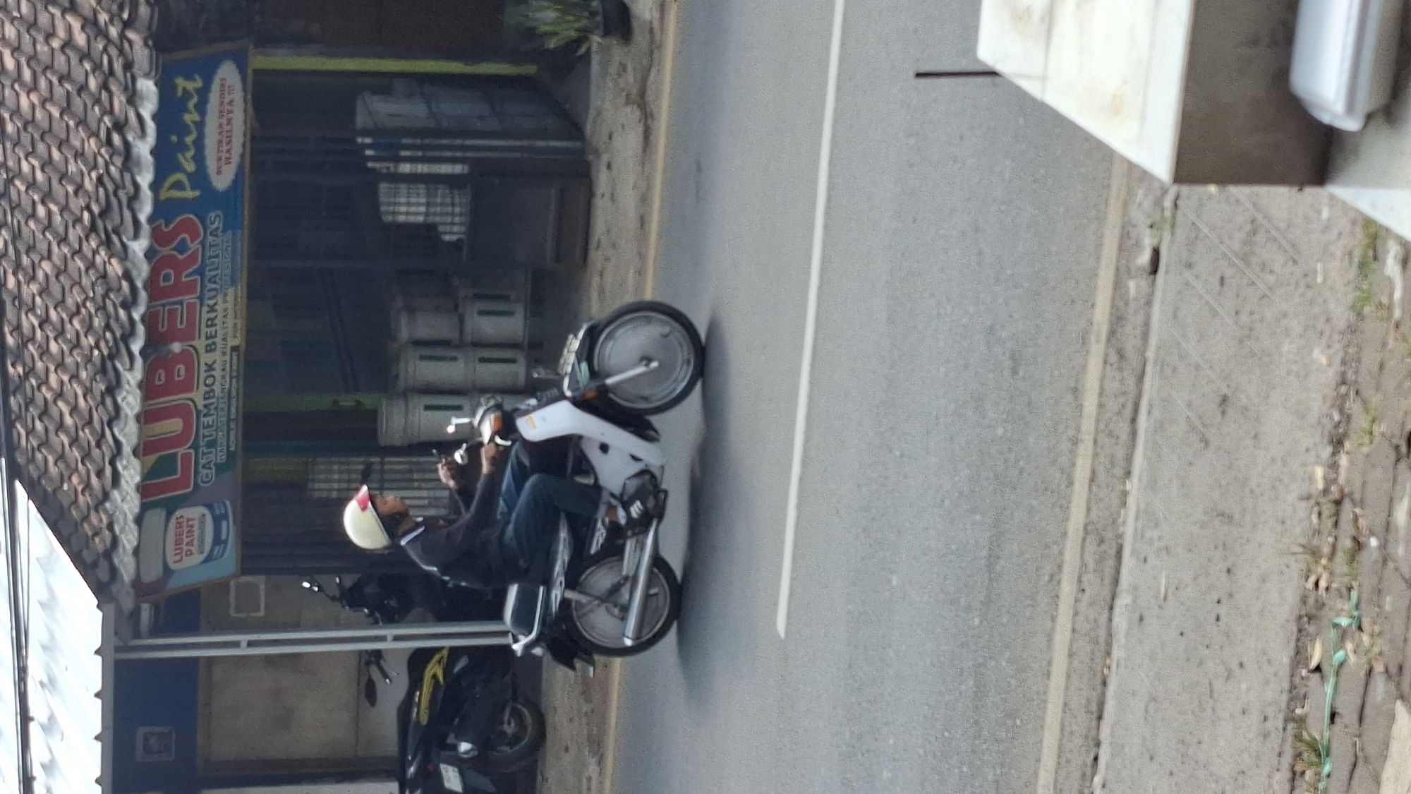 A side view captures a person riding a motorcycle on a paved road in Cilaku, Indonesia. The rider, wearing a dark long-sleeved shirt, blue jeans, and a white and red helmet, is positioned on a white and black motorcycle. The motorcycle has a silver exhaust pipe and silver wheel rims with spokes. In the background, a building with a tiled roof and a blue awning displays the sign "LUBERS PAINT" with the tagline "CAT TEMBOK BERKUALITAS" (Quality Wall Paint). Several white paint cans are stacked near the building entrance. The road is marked with a white line. The environment appears to be a commercial area with some greenery visible.
