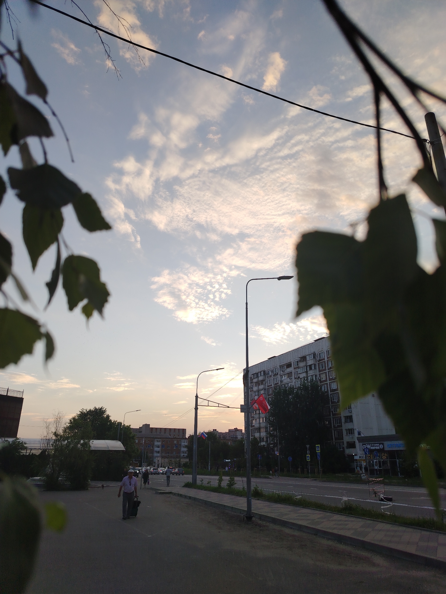 The image depicts a street scene in Krasnodar, Russia, at dusk. In the foreground, blurred green leaves frame the view. A man is walking across a paved area, pulling a dark suitcase on wheels. Behind him, a low-rise building with a white awning is visible, surrounded by trees. Further in the background, a tall apartment building with numerous windows dominates the skyline. A street with traffic and a sidewalk runs parallel to the apartment building. Streetlights are present, and a Russian flag is affixed to a pole. The sky is a pale blue with scattered, wispy clouds, illuminated by the setting sun. The overall atmosphere is that of an urban evening.