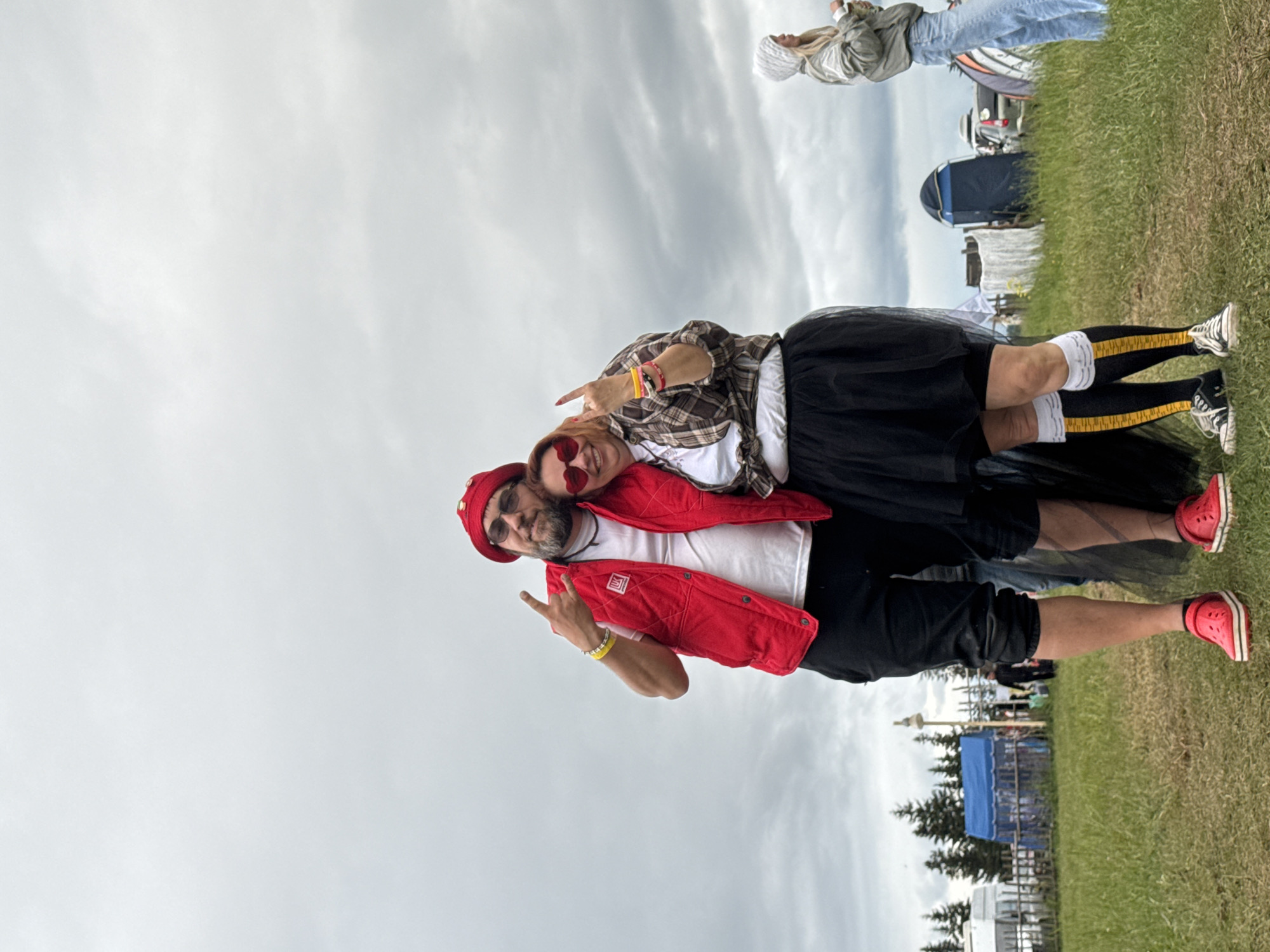 An outdoor scene in Bol'shaya Kivara, Russia, depicts two individuals posing for a photograph in a grassy field under an overcast sky. The male on the left, with a beard and sunglasses, wears a red knit beanie with embellishments, a red quilted vest with a white "LUK" logo over a white t-shirt, black cropped pants, and red casual shoes. He has a yellow wristband and makes a "V" sign with his left hand while embracing the woman next to him. The female on the right, with reddish hair, wears red heart-shaped sunglasses, a brown and white plaid shirt tied over a white t-shirt, a black tulle skirt, black knee-high socks with yellow and white stripes, and black and white low-top sneakers. She has multiple colorful wristbands and makes a "rock and roll" hand gesture with her right hand. Both individuals are smiling.
In the background, from left to right, are evergreen trees, a white van, a wooden fence, a blue pop-up tent, a tall light-colored pole, a white tarp structure, a dark blue portable tent (possibly a toilet or changing room), and other vehicles and tents further in the distance. Another person in a white hooded jacket and blue jeans stands to the far right. The ground is covered in green and dry grass.