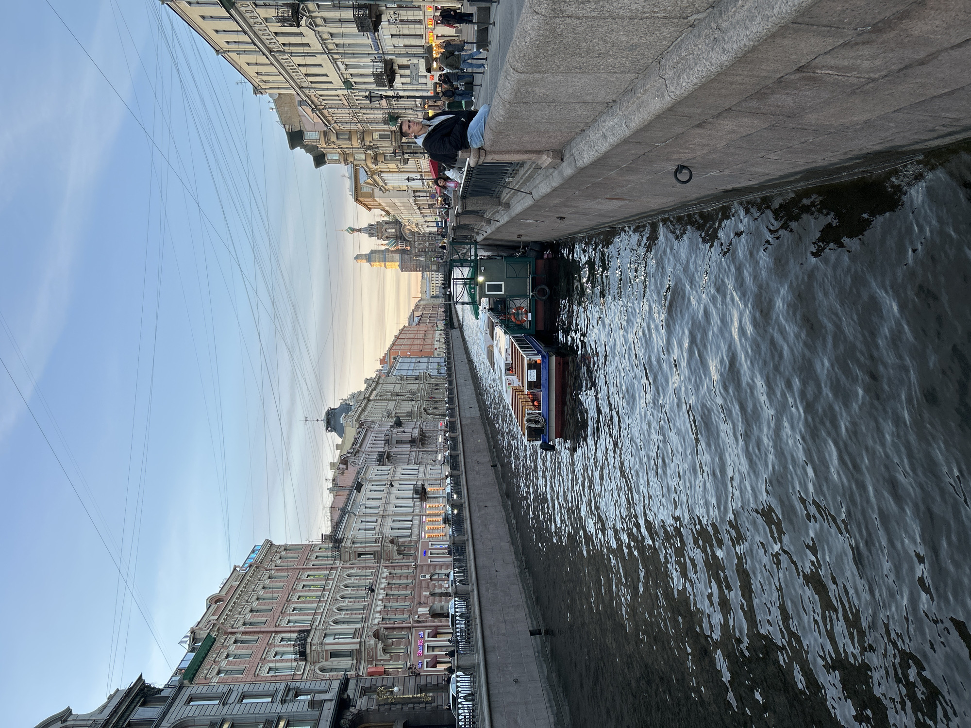 A panoramic view captures a canal scene in Saint Petersburg, Russia, during what appears to be late afternoon or early evening. The image is composed of a dark, rippling canal in the foreground, flanked by stone embankments and historic buildings under a clear, pale blue sky.
On the right side of the frame, a grey stone embankment separates a pedestrian walkway from the canal. A person wearing a black jacket, blue jeans, and light-colored shoes is seated on the edge of this embankment, facing across the canal. Further down the walkway, other people are visible, though less distinctly. A black metal mooring ring is attached to the inner wall of the embankment near the water level.
Across the canal, on the left side, an uninterrupted row of ornate, multi-story buildings with classical architectural details lines the waterway. These buildings feature numerous windows and facades in various pastel shades of beige, pink, and grey. Some windows and signs on these buildings emit a warm, artificial glow, contrasting with the fading natural light. On one building, a vertical sign is partially visible, reading "BRONNITSY" (likely "БРОННИЦЫ" or "Бронницкий" in Russian), with a horizontal sign below it appearing to say "CAFE" (likely "КОФЕ" in Russian).
Several boats navigate or are moored in the canal; one prominent boat has a distinctive green cabin. Above the buildings, numerous thin, dark overhead wires crisscross the sky, adding an urban texture to the serene backdrop. The sky exhibits a gentle gradient from light blue to a warmer, lighter hue near the horizon, indicative of the time of day. The overall atmosphere is calm, depicting typical urban life along the historic waterways of Saint Petersburg.