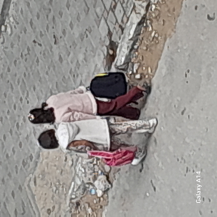 Two individuals, appearing to be young, walk away from the camera on a grey, textured ground in Khan Yunis, Palestinian Territory. The person on the left wears a white vest over patterned clothing and white sneakers, carrying a pink backpack in their right hand. The person on the right wears a light pink top and dark red pants, carrying a dark blue/black backpack with a yellow accent in their left hand. To their right, a damaged concrete curb separates the walking surface from a higher, lighter grey paved area with a block pattern. Debris, including small rocks and litter, is visible along the curb. The image contains a "Galaxy A14" watermark in the bottom-left corner.