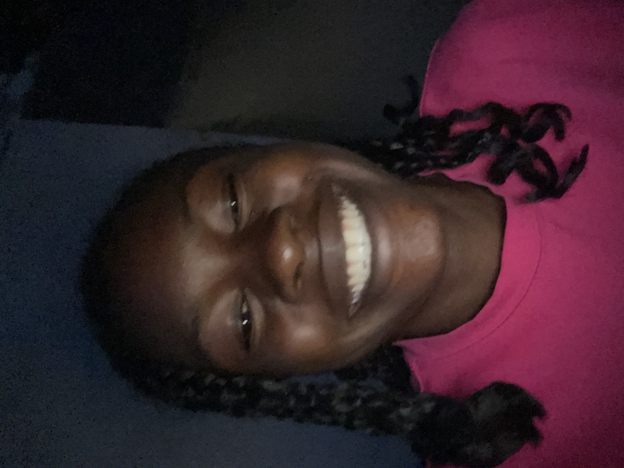 The image is a close-up selfie of a young Black woman with dark skin and braided hair, wearing a bright pink t-shirt. She is smiling widely, showing her teeth. The lighting is dim, suggesting the photo was taken indoors in low light conditions, possibly at night. The background is dark and out of focus, with a hint of a blueish-gray wall on the left. Her hair is styled in two braids that fall forward over her shoulders. The image captures a candid moment of joy.