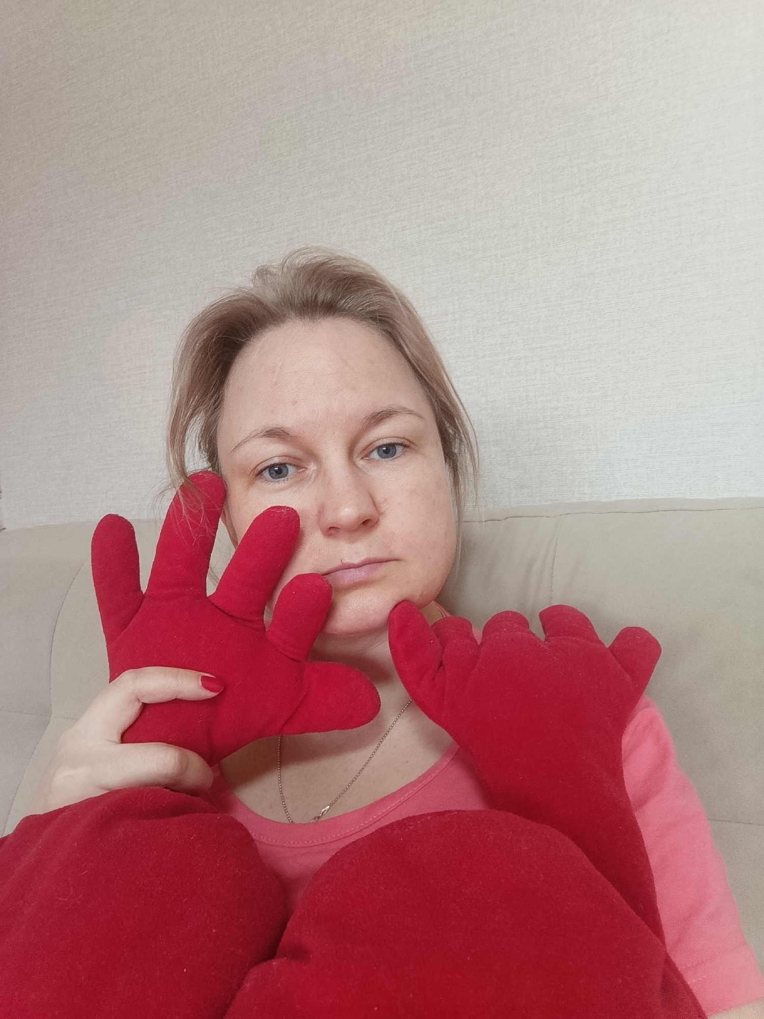 A woman with fair skin and light blonde hair is holding two red, five-fingered plush hands up to her face, one on either side. She is wearing a light pink t-shirt and a delicate silver necklace. She appears to be seated on a beige upholstered couch. The background is a textured beige wall. The location is identified as Марковский, Russia, but no elements in the image confirm this.