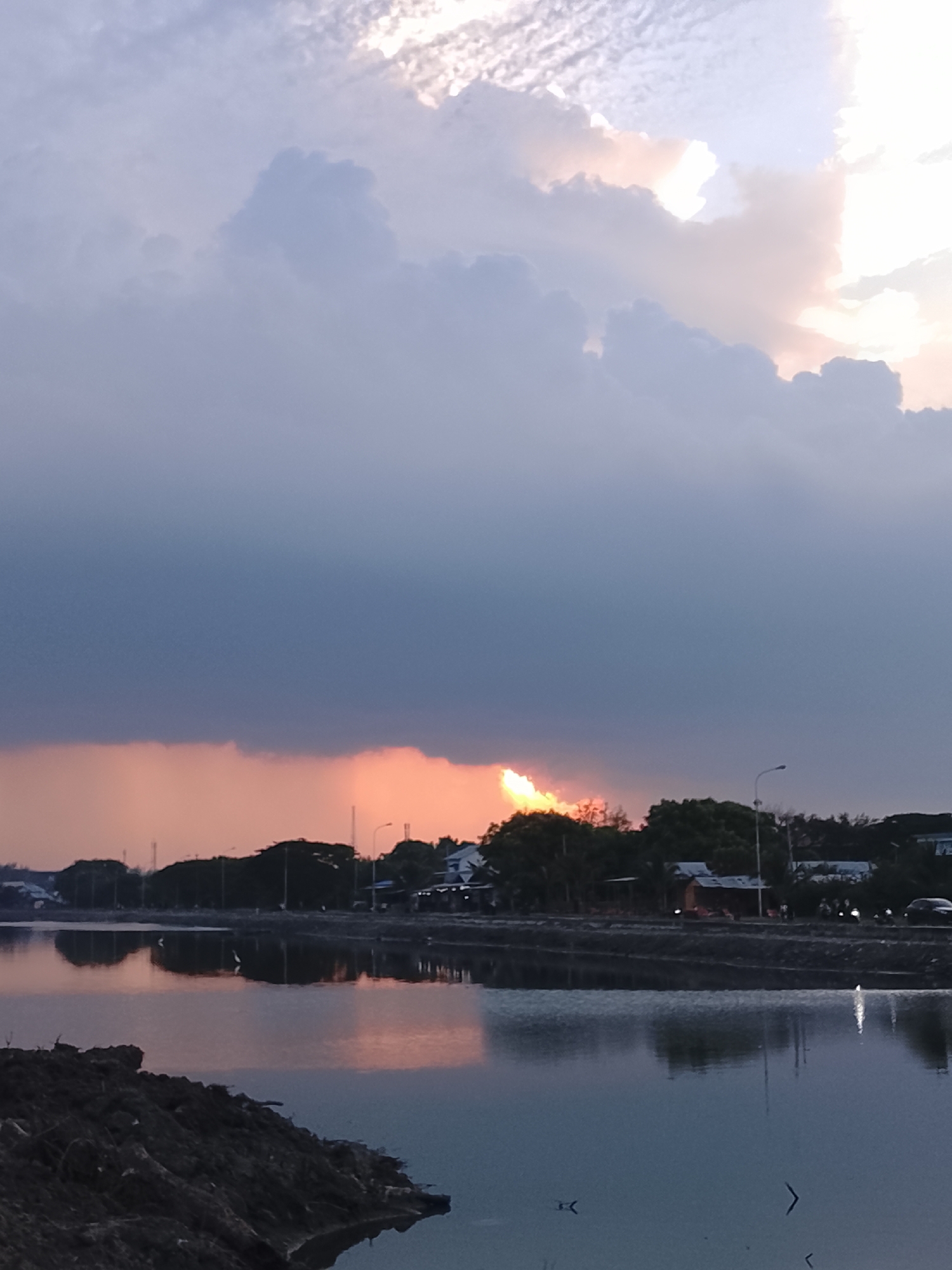 The image depicts a sunset over a body of water, likely a pond or fish farm, in Lhokseumawe, Indonesia. The sky is filled with dramatic clouds, with the sun's rays breaking through, casting an orange glow. In the distance, a treeline with scattered buildings and streetlights is visible along the horizon. The water reflects the colors of the sky and the silhouettes of the trees and structures. A pile of dark soil is in the foreground on the left side of the image. There are no identifiable people clearly visible, but the presence of streetlights and buildings suggests an inhabited area. The overall scene conveys a tranquil and atmospheric twilight ambiance.