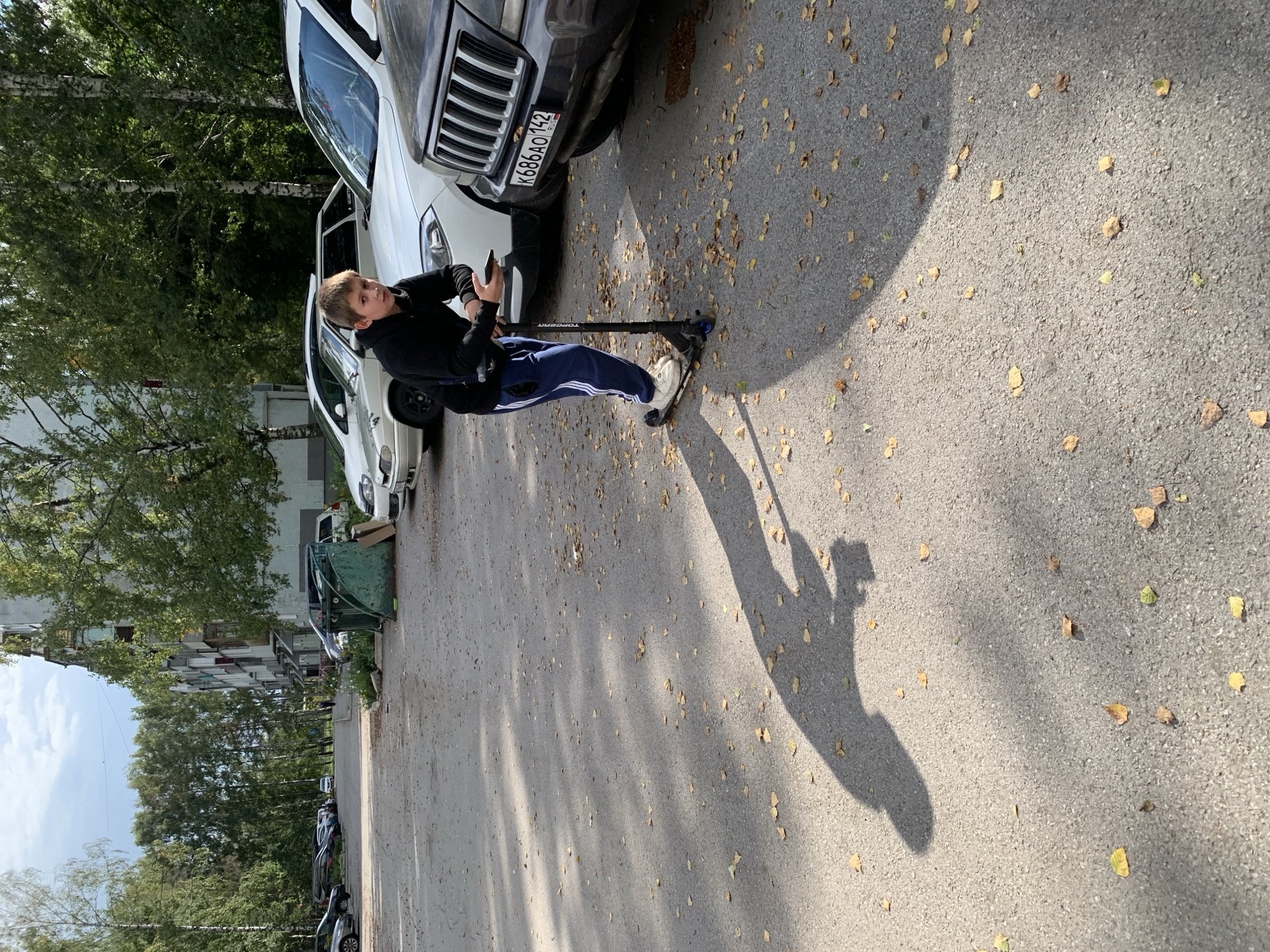 A teenage boy in a black hoodie and blue track pants with white stripes is standing on a black scooter on an asphalt parking lot. He is holding a smartphone in his right hand and appears to be looking at it. The scooter is dark colored with blue accents. In the background, several cars are parked, including a dark colored Jeep in the foreground with a Russian license plate K686AO 142. To the left of the Jeep is a white SUV. Further in the background, a light-colored sedan is parked, and behind it, a row of trees line the parking lot. A multi-story apartment building with balconies is visible behind the trees. The ground is covered with scattered fallen leaves. The scene appears to be in a residential area in the city of Prokopyevsk, Russia, based on the license plate and the typical Soviet-era apartment building architecture. The lighting suggests it is daytime, with shadows cast by the trees and the boy on the scooter.