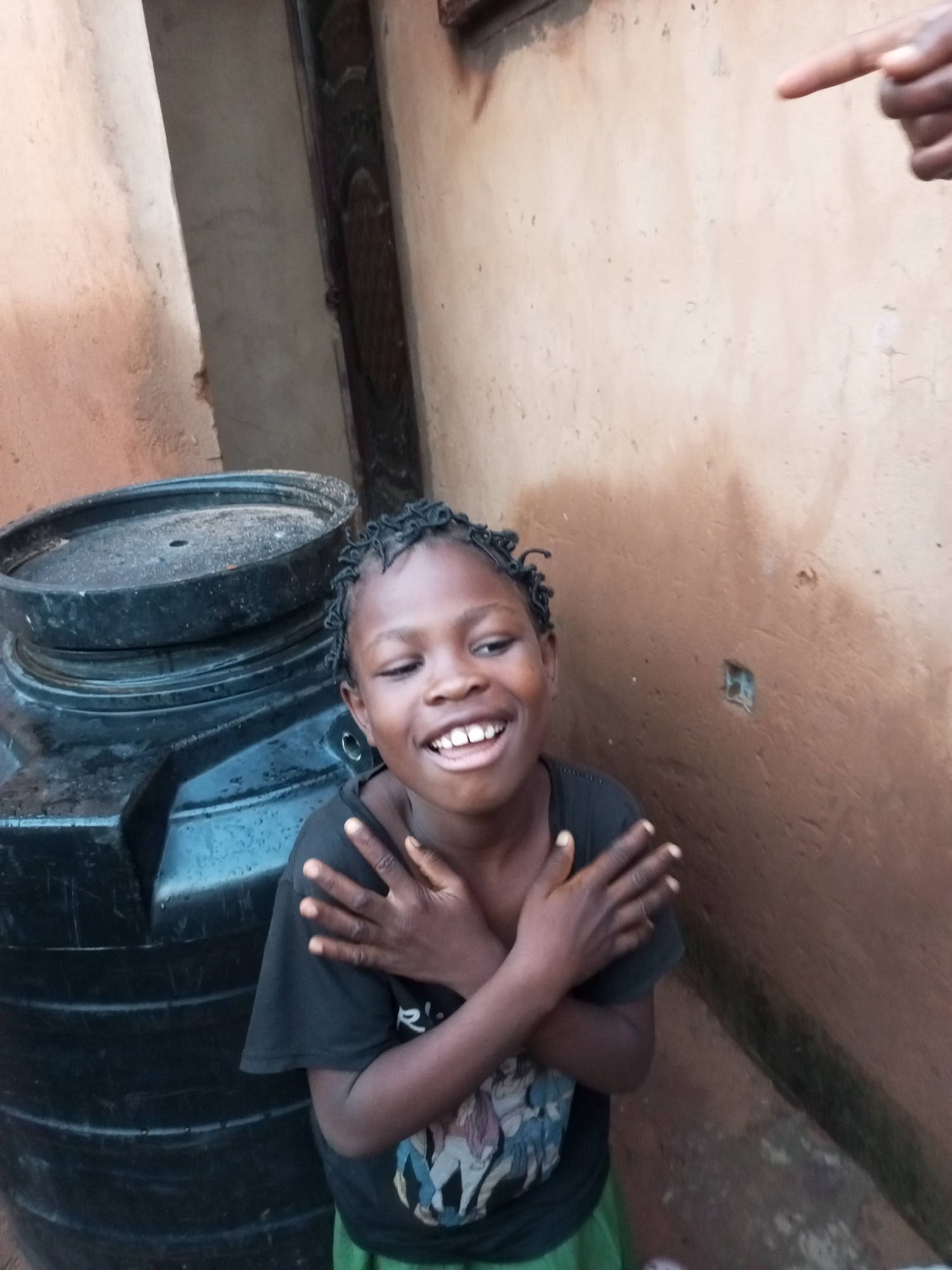 In the city of Tempure, Nigeria, a young Black child with braided hair smiles widely, their hands crossed over their chest. The child wears a black t-shirt with a graphic print and a green skirt. They are positioned next to a large black water tank, which is partially visible on the left side of the frame. The background consists of a textured, light brown wall that appears to be made of mud or stucco. A small, dark rectangular opening is visible in the wall behind the child. A hand is seen in the upper right corner of the image, with a finger pointed towards the scene. The lighting suggests an outdoor setting during the day.