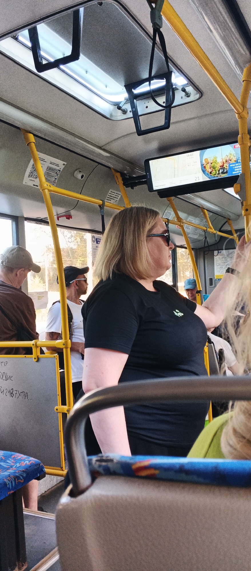 The image depicts the interior of a public bus in Izhevsk, Russia. The primary subject is a woman with blonde hair and sunglasses, wearing a black t-shirt. She is standing and appears to be holding onto a yellow pole. In the foreground, a portion of a gray seat and a blue patterned fabric seat are visible. Other passengers can be seen in the background, including a man wearing a cap and a man in a white t-shirt and black cap. A digital screen displaying information and an image is mounted above the passengers. Yellow poles and handrails are a prominent feature of the bus interior. The lighting suggests it is daytime.