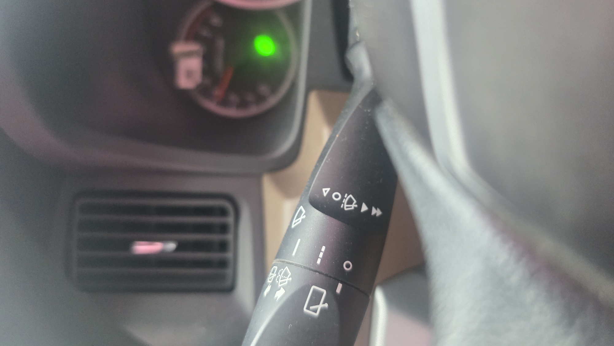 The image shows the interior of a vehicle. A black wiper/washer stalk is visible to the right, displaying various icons related to wiper functions. Behind the stalk, a portion of the dashboard is seen, featuring an air vent with horizontal slats and a gauge with a green light illuminated. The exact location in Teupinbatee, Indonesia, is not discernable from the given visual data.