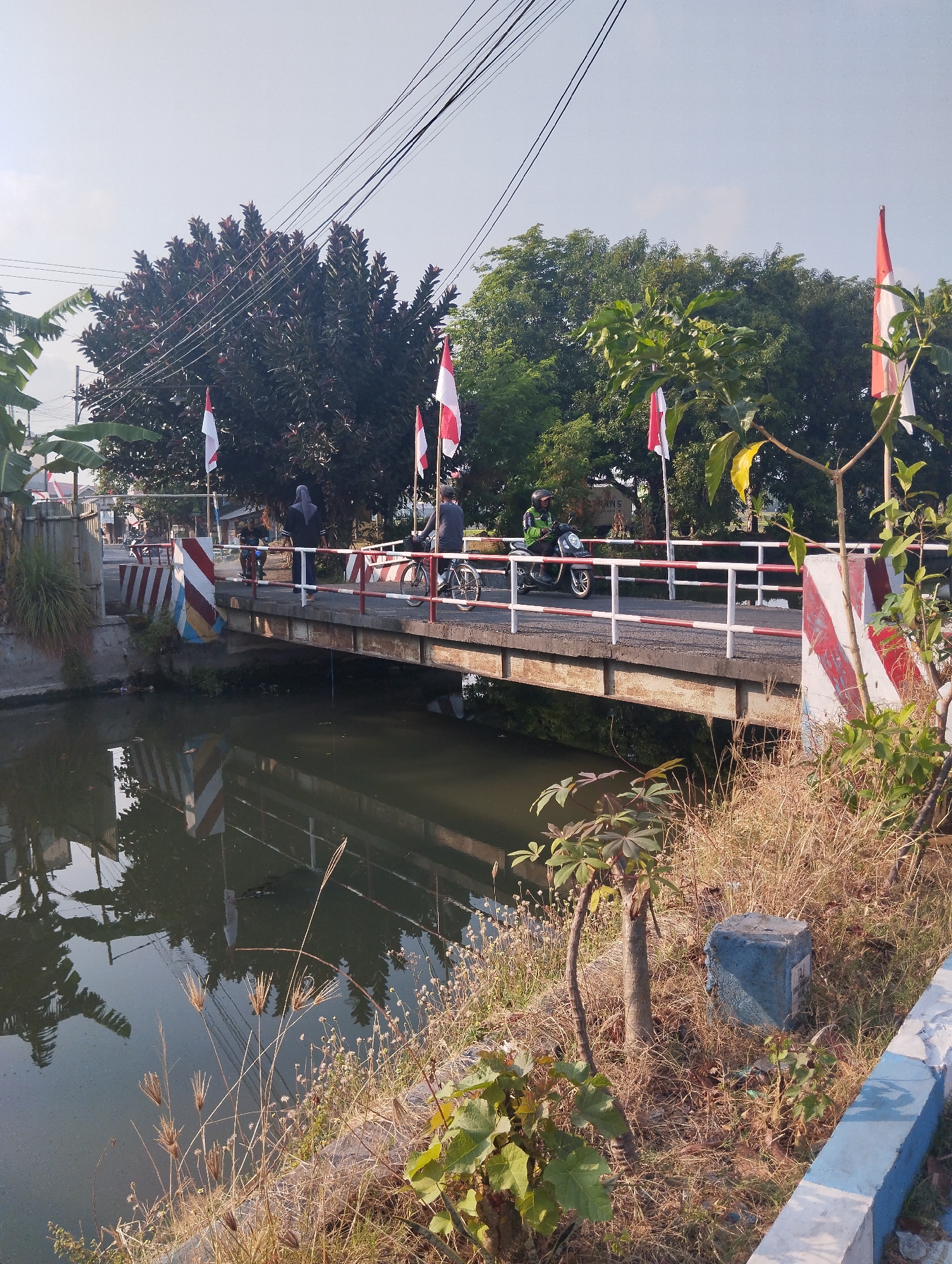 The image shows a bridge over a canal in the city of Sawotratap, Indonesia. The bridge has a metal railing with red and white stripes. There are several Indonesian flags displayed on poles along the bridge. Two people are on the bridge, one riding a motorcycle and another walking. The canal water is dark and reflects the bridge and surrounding trees. The banks of the canal are overgrown with dry grass and some small plants. In the background, there are trees and power lines. The sky is clear and blue.