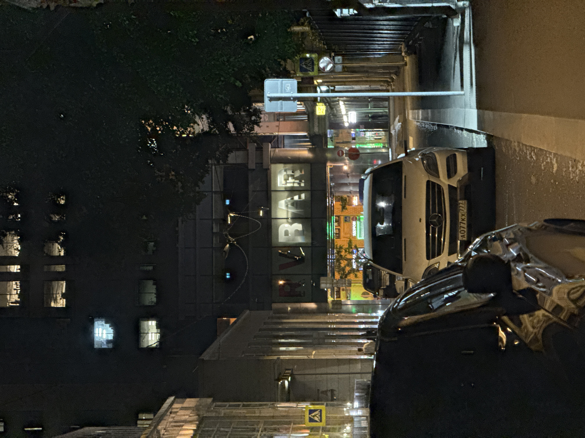 This nighttime photograph captures a wet urban street scene in Moscow, Russia. The street, slick with rain or recent rainfall, reflects the numerous artificial lights from buildings and street lamps, creating a luminous effect across the pavement.
On the right side of the street, several vehicles are parked. Most prominently, a white Mercedes-Benz Sprinter van faces towards the viewer, its front grille and headlights clearly visible. Its Russian license plate, "A077KX 777", indicates its origin in the Moscow region. In front of the van, a dark-colored car, possibly a sedan, is partially visible, showing its side mirror and rear section.
To the left, dark foliage of trees partially obscures some buildings. One building, central in the frame, features large, illuminated white letters on its facade that appear to spell "BAF" or potentially "BAR". Other multi-story buildings line the street, some with lit windows contributing to the ambient glow. Further down the street, reflections of signs and indistinct shopfronts can be seen on the wet ground.
Street furniture includes a pole with a blue rectangular sign (possibly a bus stop or parking indicator), a convex mirror for visibility, and a yellow diamond-shaped pedestrian crossing sign featuring a white downward arrow. No people are visible, and the scene depicts a quiet, urban environment after dark, suggesting recent rainfall.