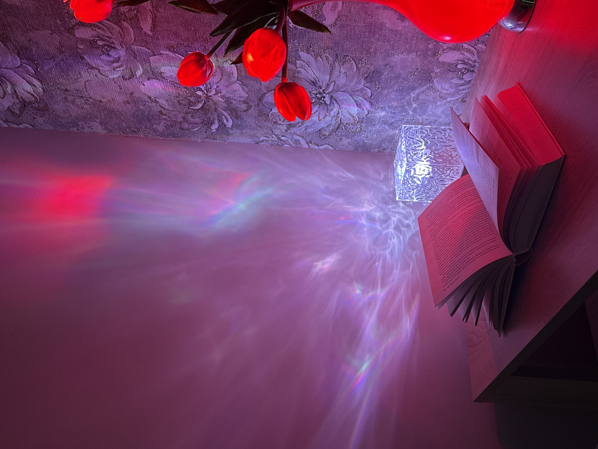 A top-down, high-angle shot reveals a corner of a room. The wall, a patterned wallpaper of flowers in shades of purple and white, is illuminated by red light from an unseen source, possibly a lamp with red tulip-shaped shades overhead. A small, square, decorative lamp is also visible. The light from the lamps creates complex patterns on the adjacent wall, casting shadows and refractions. A partly open book rests on the edge of a dark wooden table. The room gives a strong domestic or interior setting. There is no known city or region apparent from the image.