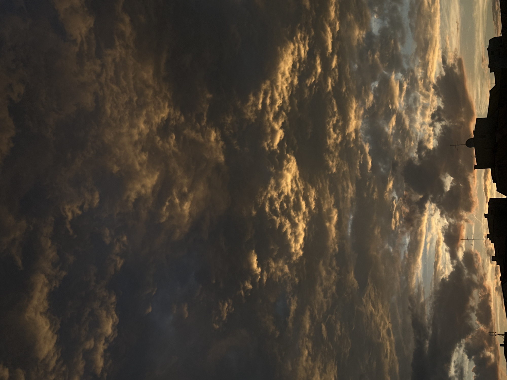 The image presents an urban landscape at either sunrise or sunset, specifically located in Sofia, Bulgaria. The upper two-thirds of the frame are dominated by a textured sky filled with dense formations of altocumulus or stratocumulus clouds. These clouds exhibit a palette of dark gray and black in their shadowed areas, intensely contrasted by patches of vibrant golden-orange and yellow illumination from a low sun, particularly in the mid-section and towards the horizon. A small, lighter area of pale blue sky is visible through a break in the clouds near the lower-middle of the image. The bottom quarter of the image reveals the dark silhouettes of several urban buildings with flat rooftops, positioned against the illuminated horizon. Two distinct antennas are visible on the rooftops: a multi-pronged structure on a central building and a smaller array to the left. No individuals or specific human activities are observable in the scene.