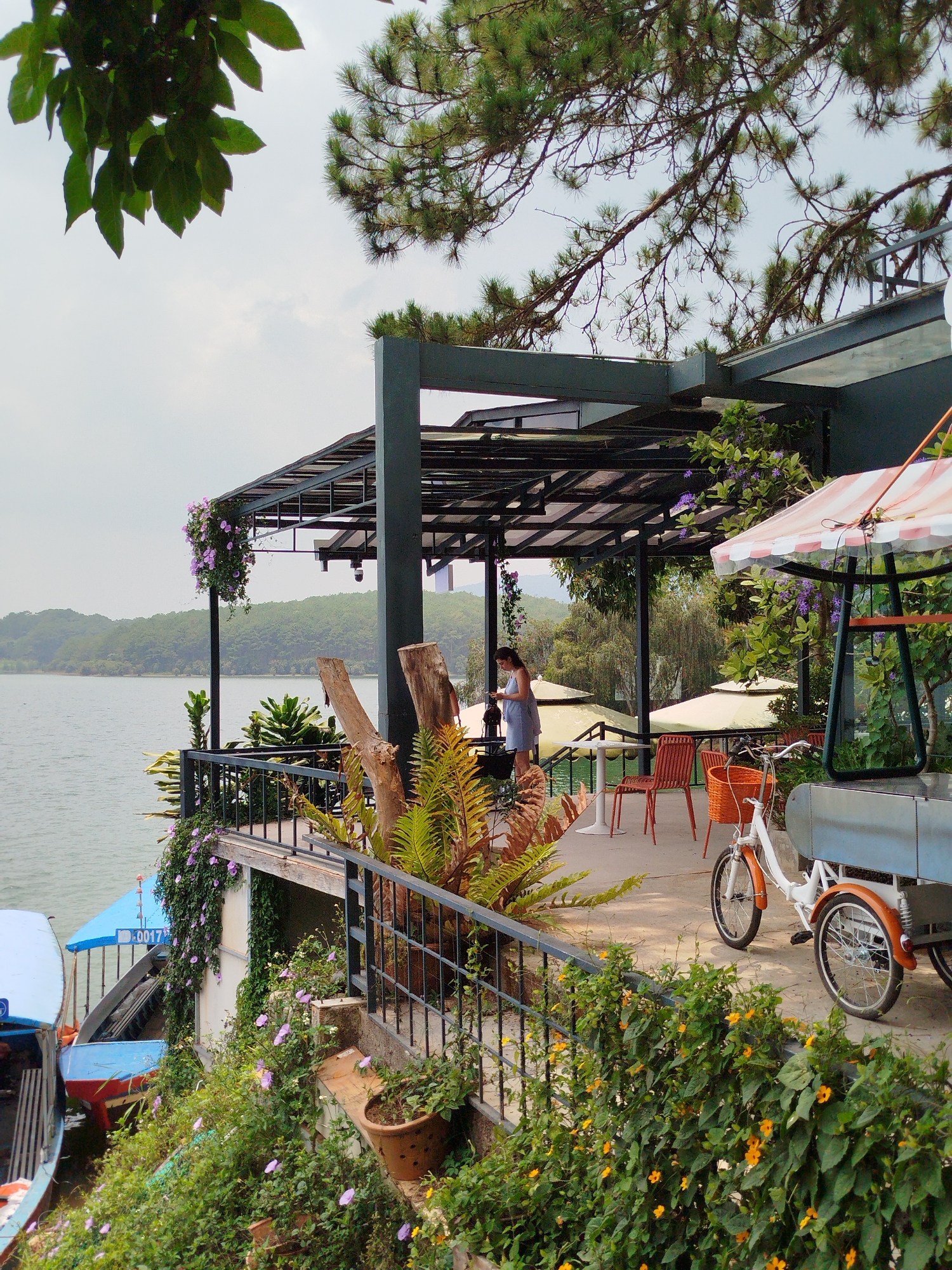 The image depicts a scenic view from a waterfront cafe in Da Lat, Vietnam. The cafe, situated on a raised platform, overlooks a tranquil lake surrounded by lush greenery.
A woman stands on the platform, facing away from the viewer. Orange chairs and tables are positioned on the patio.
A three-wheeled bicycle with a food cart is parked near the edge of the platform. A striped umbrella is affixed to the cart. The platform is decorated with vibrant flowers and plants cascading over the railing.
Below the platform, several boats are docked, adding to the ambiance of the lakeside setting. The overall impression is one of a relaxed and picturesque destination.