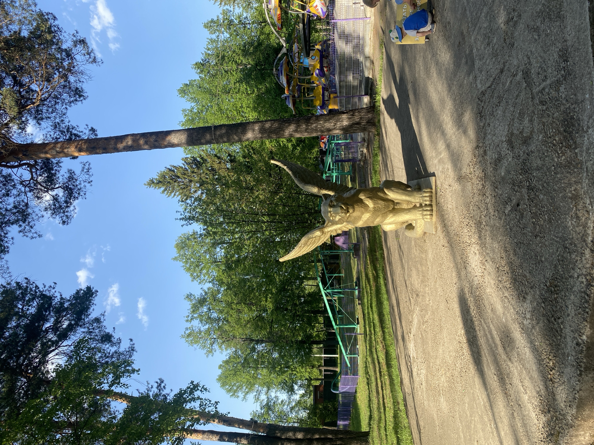 The image depicts a sunny day in a park in Izhevsk, Russia. A golden-colored griffin statue stands on a paved path. In the background, green trees and a portion of a colorful amusement park ride are visible. A tall tree trunk stretches across the scene from the left, partially obscuring the sky. Another, smaller tree is on the left, while more trees are clustered in the background. A small child in a blue shirt and white shorts kneels on the pavement near the statue, appearing to interact with something on the ground. Grass and park equipment are also visible in the background.