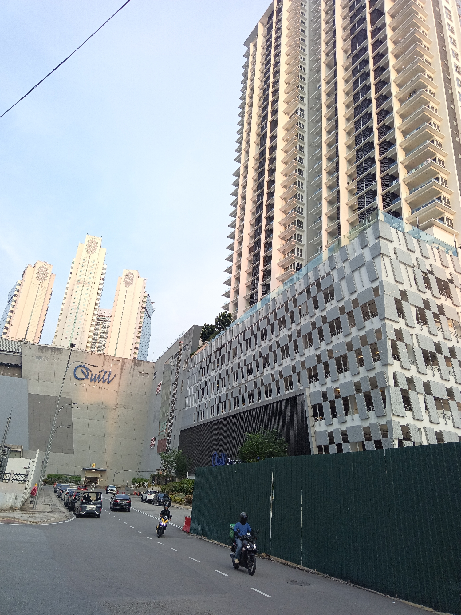 In the city of Kuala Lumpur, Malaysia, a street scene unfolds with modern architecture. Dominating the background are several tall, cream-colored residential buildings with numerous balconies. In the mid-ground, a complex structure with a grey, patterned facade, likely a car park, is attached to a large, grey concrete building bearing the blue logo "Quill City Mall." In the foreground, a two-lane road curves to the right. Several cars are parked along the left side of the road and a few are in motion. A motorcycle with a rider wearing a blue shirt and a helmet is traveling on the right lane, moving away from the camera. Another motorcycle with a rider in black is visible further down the road. A dark green corrugated metal fence runs along the right side of the street. The sky is a pale blue, suggesting it is either early morning or late afternoon.