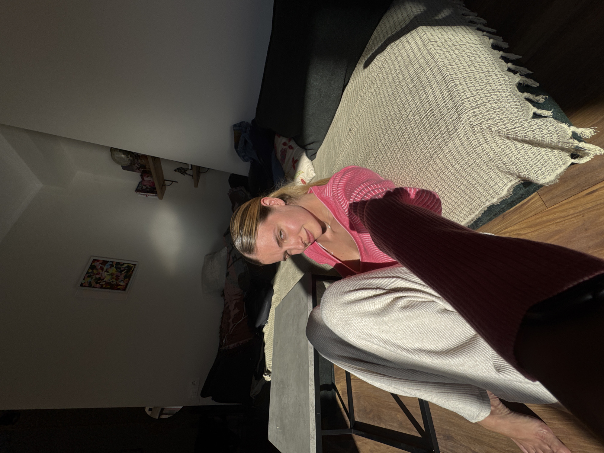 A low-angle, tilted image captures an indoor scene, primarily featuring a woman looking towards the camera. She is positioned on the left side of the frame, with her head slightly tilted and a subtle smile. Her light brown hair is pulled back, and she is wearing a pink, ribbed long-sleeved top and light beige, wide-leg pants. Her right bare foot is visible in the bottom right corner, resting on a wooden floor. She appears to be sitting on a low stool or chair, with a light gray, rectangular table or surface in front of her.
The setting is an interior room, likely a living area. To the right of the woman, a textured, light beige blanket with tassels is draped over what appears to be a sofa, which is covered with dark fabric. The room is dimly lit, with a strong, directional light source (possibly sunlight or a bright lamp) creating highlights on the woman's face, clothing, and the blanket, while casting deep shadows elsewhere. In the background to the left, a white wall features a small, framed piece of colorful abstract art and some shelving with indistinct objects. The image is taken at an angle, giving it a dynamic, informal feel. No discernible text is visible.
