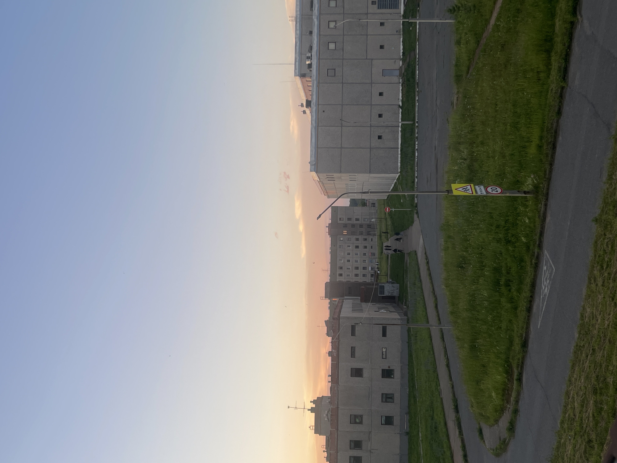 The image depicts a view of buildings and a street in the city of Markovsky, Russia, during what appears to be sunset or early evening. The sky is a gradient of pale blue at the top transitioning to soft pink and orange hues near the horizon, indicating a sunset.
In the foreground, a grey asphalt road curves through a grassy area. There are road markings on the asphalt, including a triangular white symbol. A street signpost is visible on the right, displaying three signs: a yellow triangular sign with two figures, a rectangular white sign with "100m" in black text, and a circular red and white sign with "20" inside.
Further back, several concrete buildings are arranged in a somewhat uniform, functional architectural style. On the left, a long, low-rise building with evenly spaced windows runs horizontally across the frame. To its right, another, slightly taller concrete building is visible. In the middle ground, a wider road or pathway leads towards a row of taller, apartment-style buildings. A group of people, appearing as small figures, are walking along this pathway. On the right side of the image, a large, rectangular concrete building with a flat roof dominates the mid-ground.
The overall environment suggests an urban or semi-urban setting, likely a residential or industrial area, characterized by functional architecture and open green spaces. The lighting suggests a time of day with soft, diffused light.