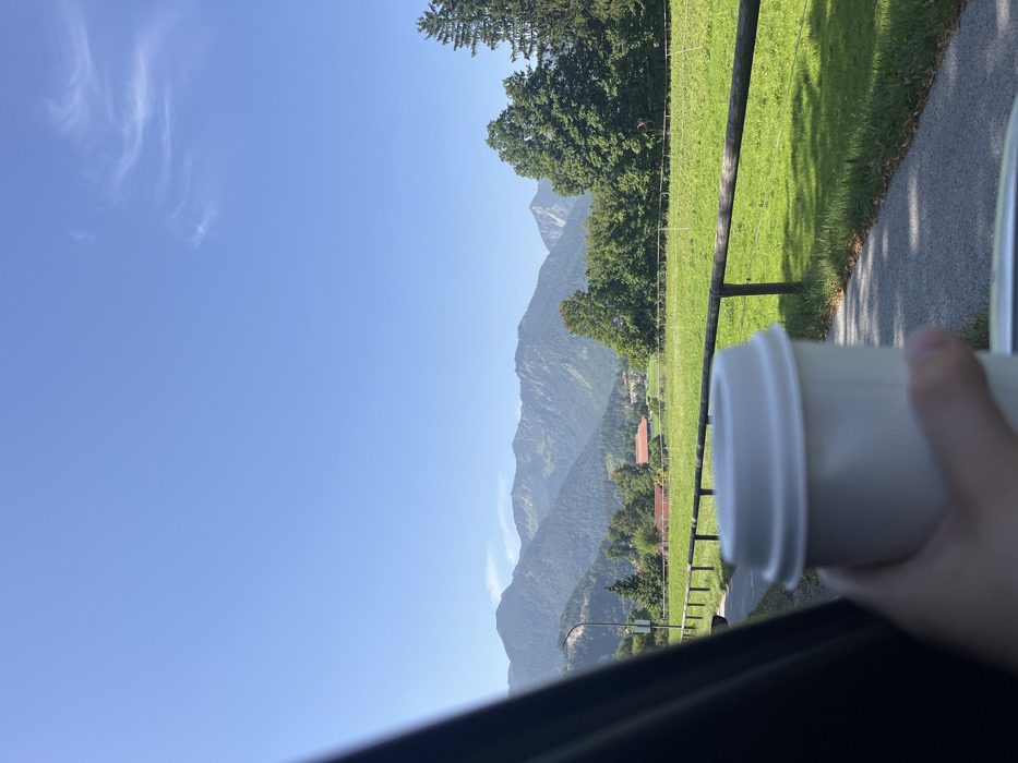 The image displays a sunny daytime scene in Fischbachau, Germany. In the immediate foreground, a human hand holds a white disposable coffee cup with a white lid, observed from within a vehicle; a dark vehicle frame is visible on the left. The midground features a paved path or road running parallel to a wooden fence, beyond which lies a large, vibrant green field. Scattered trees, including both deciduous and coniferous types, are visible behind the field. Several buildings with reddish-brown roofs are discernible in the distance, indicating a small settlement. A utility pole stands on the left side of the path. The background is dominated by a range of tree-covered mountains under a clear, bright blue sky with minimal, wispy white cloud formations. The scene is well-lit by natural sunlight, casting visible shadows.