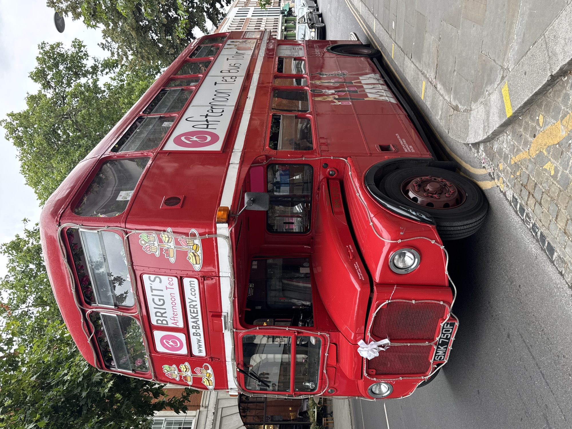 This media file shows a bright red double-decker bus, likely operating as an "Afternoon Tea Bus Tour." The bus is parked on a city street, indicated by the cobblestone and paved road, yellow double lines, and a sidewalk with buildings in the background. Trees with green foliage overhang the bus, suggesting a pleasant, possibly spring or summer, day.
The bus is decorated with branding for "BRIGIT'S Afternoon Tea" and "www.B-BAKERY.com," with illustrations of tea sets and people enjoying afternoon tea. A license plate reading "SMK 750F" is visible. A small white bow is tied to the front of the bus, hinting at a special occasion, perhaps a celebration or a wedding. The overall atmosphere appears cheerful and inviting, suggesting a tourist attraction or a mobile catering service. The image is taken from a slightly low angle, emphasizing the size and grandeur of the iconic London bus. The time of day is not explicitly indicated, but the natural light suggests it's daytime.