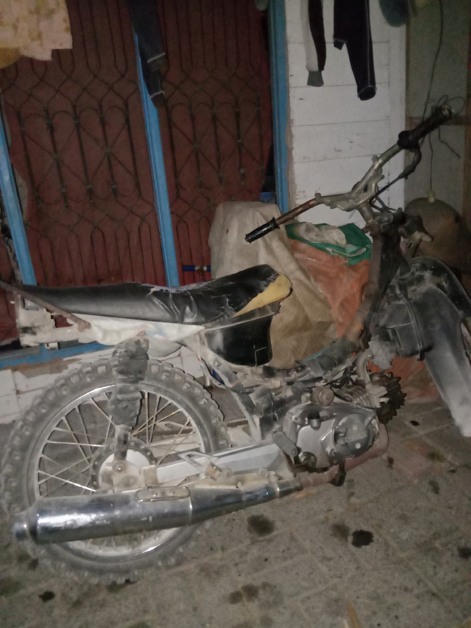 The image shows a dirt bike parked on a tiled floor, likely outdoors, with a building and a wire fence in the background. The dirt bike is black and silver, with a worn seat and a dirty exterior. The building in the background appears to be made of wood and has a red-colored door or gate with a decorative metal grill. Clothes are hanging on a wall to the right of the door. The ground is scattered with dark spots, possibly oil stains. The lighting suggests it is either nighttime or a dimly lit area. The location is identified as Jumput, Indonesia. There are no people visible in the image.