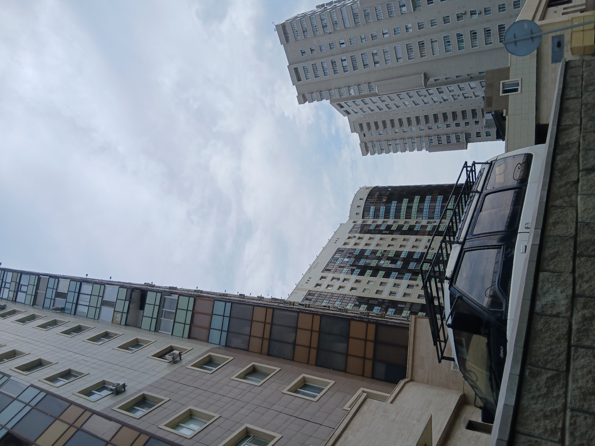 The image shows a view of several modern apartment buildings in Astana, Kazakhstan. In the foreground, the top portion of a white SUV with a roof rack is visible, parked behind a low stone wall. The buildings are tall and feature a mix of beige, brown, and glass facades with numerous windows. The sky is overcast with scattered clouds. The composition is dominated by the vertical lines of the architecture.