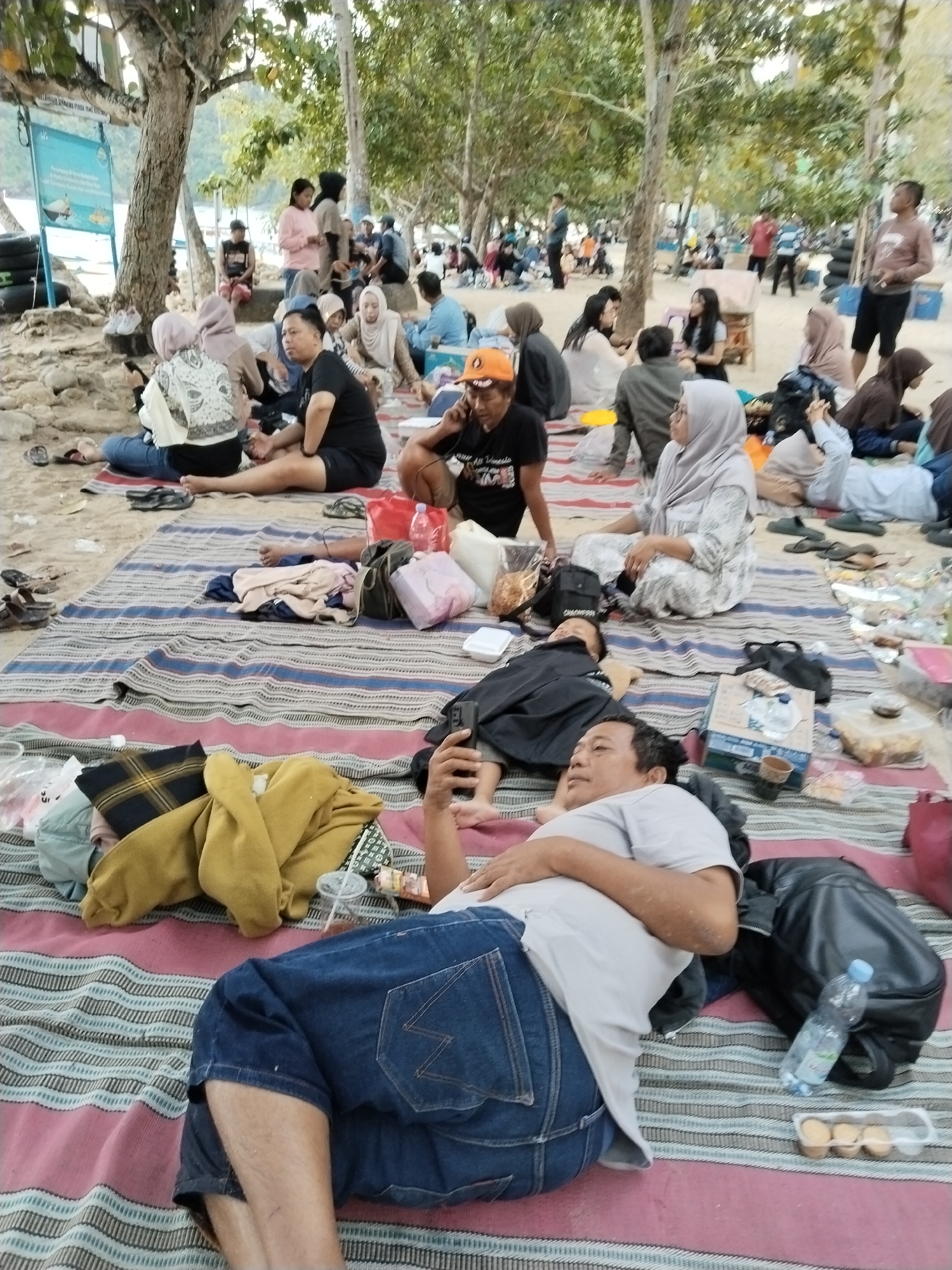 The image depicts a casual gathering of people on a beach in Prigi, Indonesia, likely during a picnic or outing. Several individuals are seen relaxing on striped mats spread on the sand. In the foreground, a man in a grey t-shirt and denim shorts is lying down, holding a smartphone and looking upwards. A black backpack and a bottle of water are nearby. To the left of the foreground man, another person is visible wearing a black t-shirt and shorts. In the middle ground, a group of people are seated on mats, some wearing headscarves. Objects like bags, clothing, and food containers are scattered on the mats. The background shows more people, trees, and a glimpse of the beach. The overall atmosphere appears relaxed and informal.