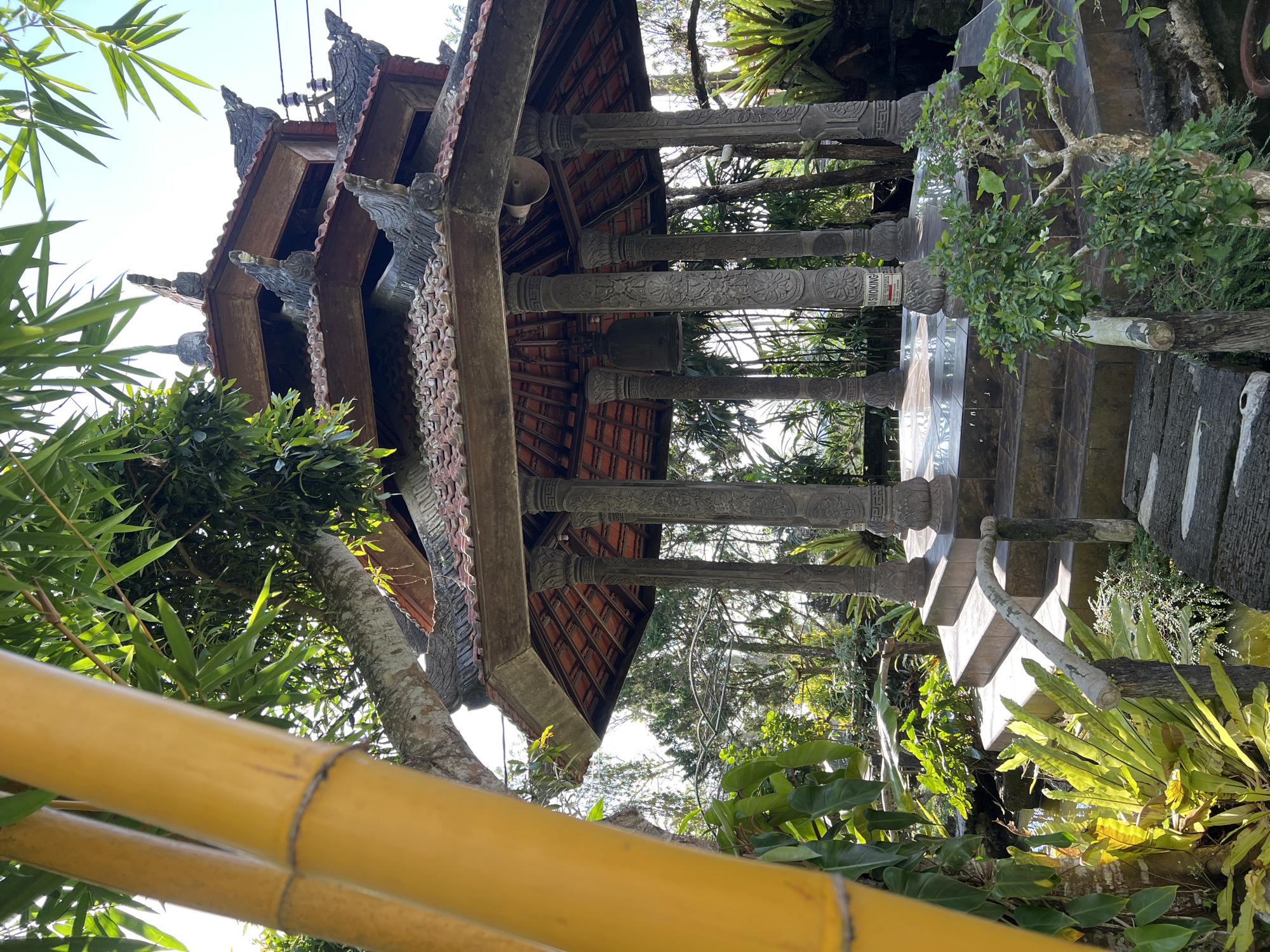 This image depicts a Balinese-style pavilion located in the city of Pupuan, Indonesia. The pavilion features a multi-tiered roof with terracotta tiles and decorative carvings on its stone pillars. A large bell hangs inside the pavilion. The structure is surrounded by lush tropical greenery, including bamboo stalks, ferns, and other plants. A small pond or water feature is visible in the foreground, and a set of stone steps leads up to the pavilion. The scene is bathed in natural sunlight, suggesting it is daytime. There are no people visible in the image.