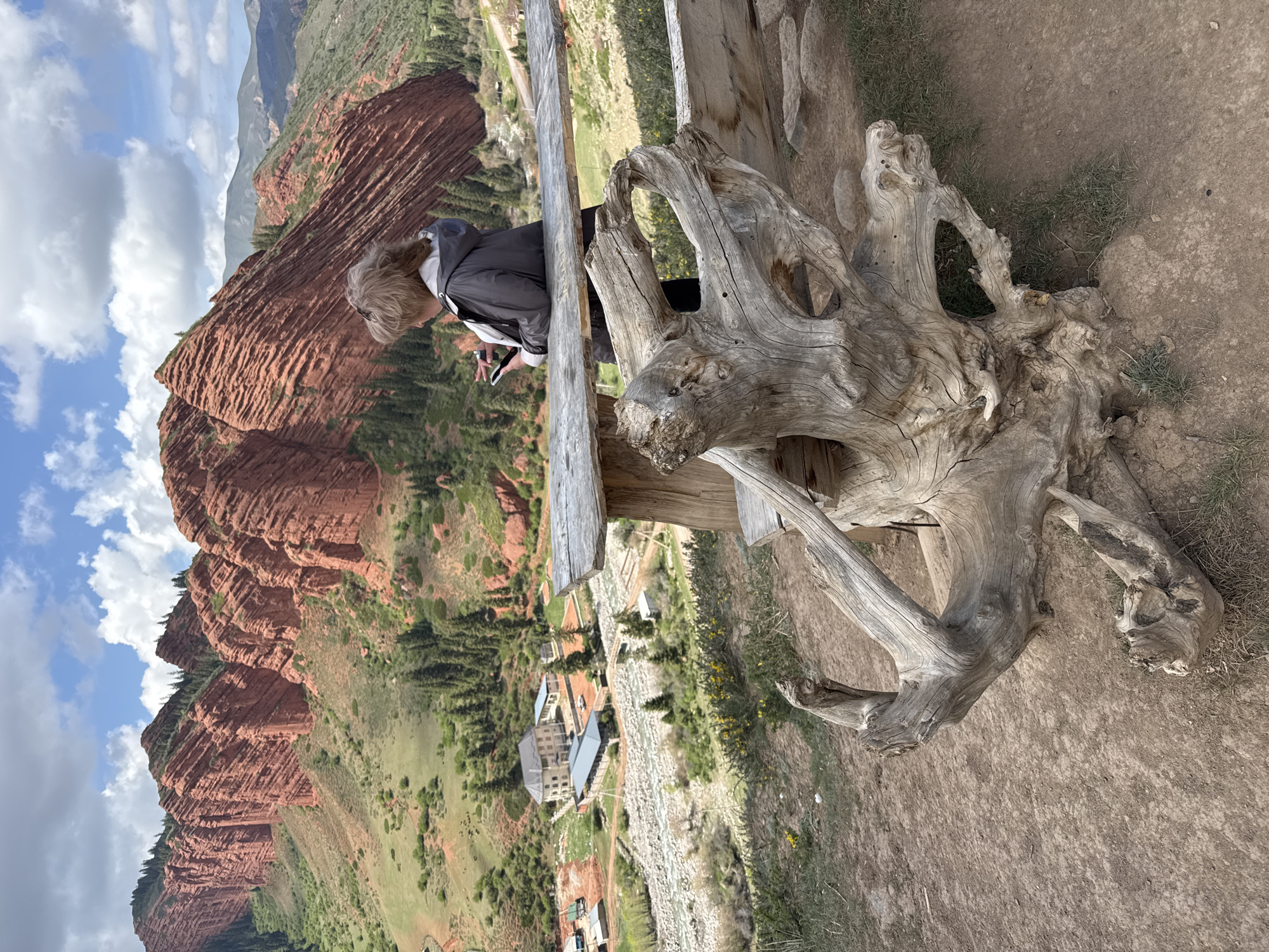 This image, captured in Jeti-Ögüz, Kyrgyzstan, presents a panoramic view of a dramatic mountainous landscape under a partly cloudy sky. On the left, towering red sandstone cliffs, characterized by horizontal layers and jagged formations, dominate the scene, interspersed with green vegetation.
In the mid-ground, a person with light-colored hair, wearing a dark jacket, stands at what appears to be a scenic overlook. Their back is partially turned to the viewer as they look down at a smartphone held in their hands, suggesting a moment of rest or observation. A rustic wooden fence or barrier stretches across the frame, providing a boundary to the viewpoint.
The foreground on the right features a large, weathered tree stump or root system with intricate, gnarled forms, adding a natural, organic element to the immediate surroundings. The ground is dry, reddish-brown earth with sparse grass.
Below the overlook, the valley floor reveals a riverbed, lush green slopes, and several buildings, indicating human presence within the vast natural environment. The bright daylight, filtered by scattered clouds, illuminates the diverse textures and colors of the landscape. The image evokes a sense of natural grandeur, offering a glimpse into the serene beauty of Jeti-Ögüz. No visible text is present.