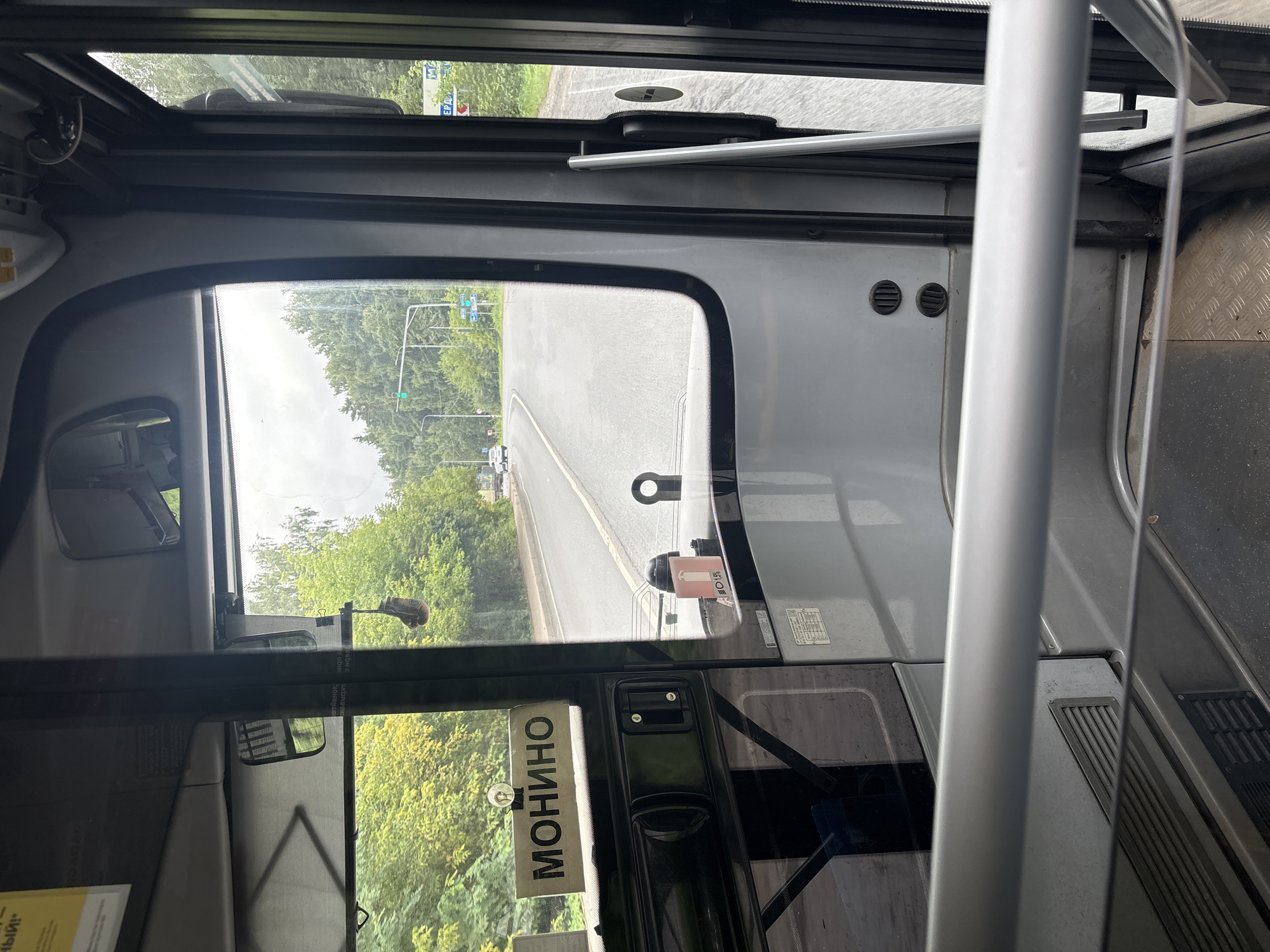 The image depicts the interior of a bus, looking forward through the windshield and side window. A sign with the Cyrillic word "МОНИНО" is visible on the dashboard. Outside, a road with trees lining both sides is seen. The road appears to be in a semi-rural or suburban area. There are traffic lights and other vehicles visible in the distance. Based on the sign "МОНИНО", this scene is likely located near Monino, which is a settlement in the Shchyolkovsky District of Moscow Oblast, Russia. The environment appears to be outdoors on a cloudy day, with lush green foliage indicating a temperate climate, likely summer or early autumn.