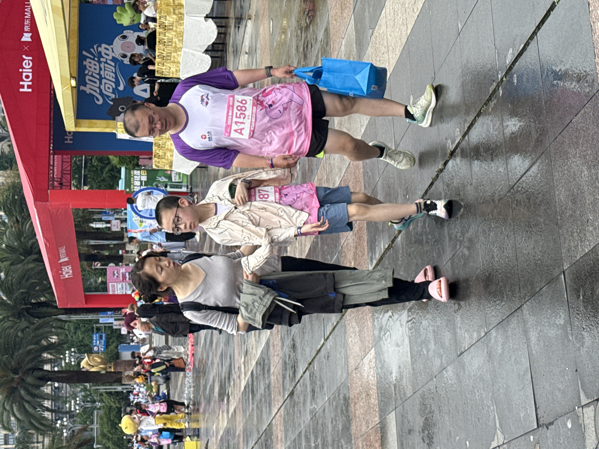The image shows a group of people walking on a paved surface in what appears to be an outdoor event setting. The location can be inferred as Nan'an District, China, due to the Chinese characters on signage, along with the "Haier" brand, a Chinese multinational company.
The paved surface is wet, suggesting recent rain or cleaning. The overall light level seems moderate, indicating daytime.
Several individuals are present. Starting from the closest, a woman with dark hair is wearing a light gray top, dark shorts, and pink sandals. Her posture and expression indicate she is walking normally.
Next to her is a younger person with glasses wearing a light-colored patterned shirt and shorts. They appear to be participating in an event, wearing a bib with the number 87.
Above them, another individual wearing glasses and a purple-sleeved shirt with a pink bib labeled "A1586" is carrying a blue bag.
In the background, a larger crowd of people is visible. A red structure with "Haier × 京东MALL" is present, along with a blue sign bearing the characters "加油!向前冲," which translates to "Add oil! Move forward!"
Overall, the scene depicts people walking in a public space, possibly after participating in or attending an event in Nan'an District, China.