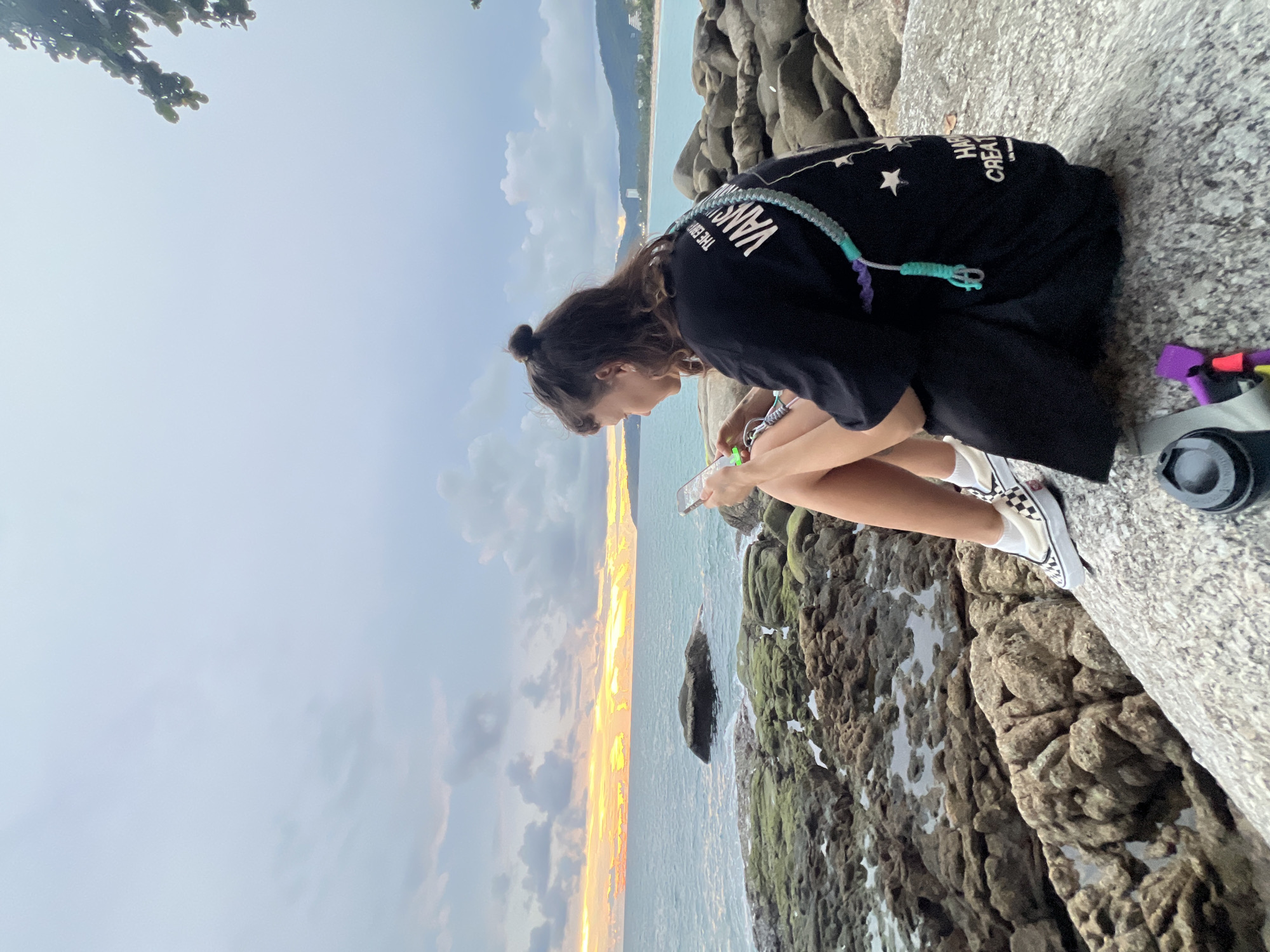 A young woman with her hair in a bun is sitting on large grey rocks on a beach, looking at her phone. She is wearing a black t-shirt with white writing and graphics on the back, and white checkered Vans sneakers with white socks. A braided strap in shades of blue and purple is draped over her shoulder. The ocean stretches out before her, with the sunset casting an orange glow on the horizon. The beach is rocky and covered in moss in some areas. In the foreground, a black thermos and a few colorful objects are visible on the rocks. The scene is set against a cloudy sky. The location is Karon Beach, Phuket, Thailand.