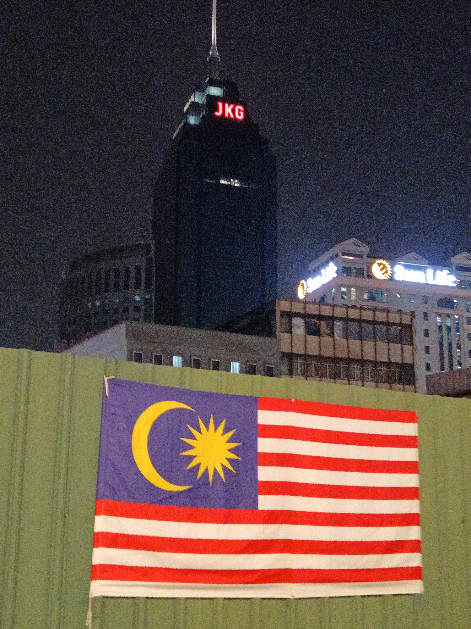 The image is a nighttime photograph taken in Kuala Lumpur, Malaysia, featuring a Malaysian flag in the foreground, partially obscuring a green corrugated metal fence. Behind the fence, several buildings are visible. The most prominent building is a dark skyscraper with red illuminated lettering that reads "JKG". To the right of the skyscraper, a lighter-colored building with illuminated signage that says "Sun Life" and features a sun logo is visible. The sky is dark, indicating it is nighttime.