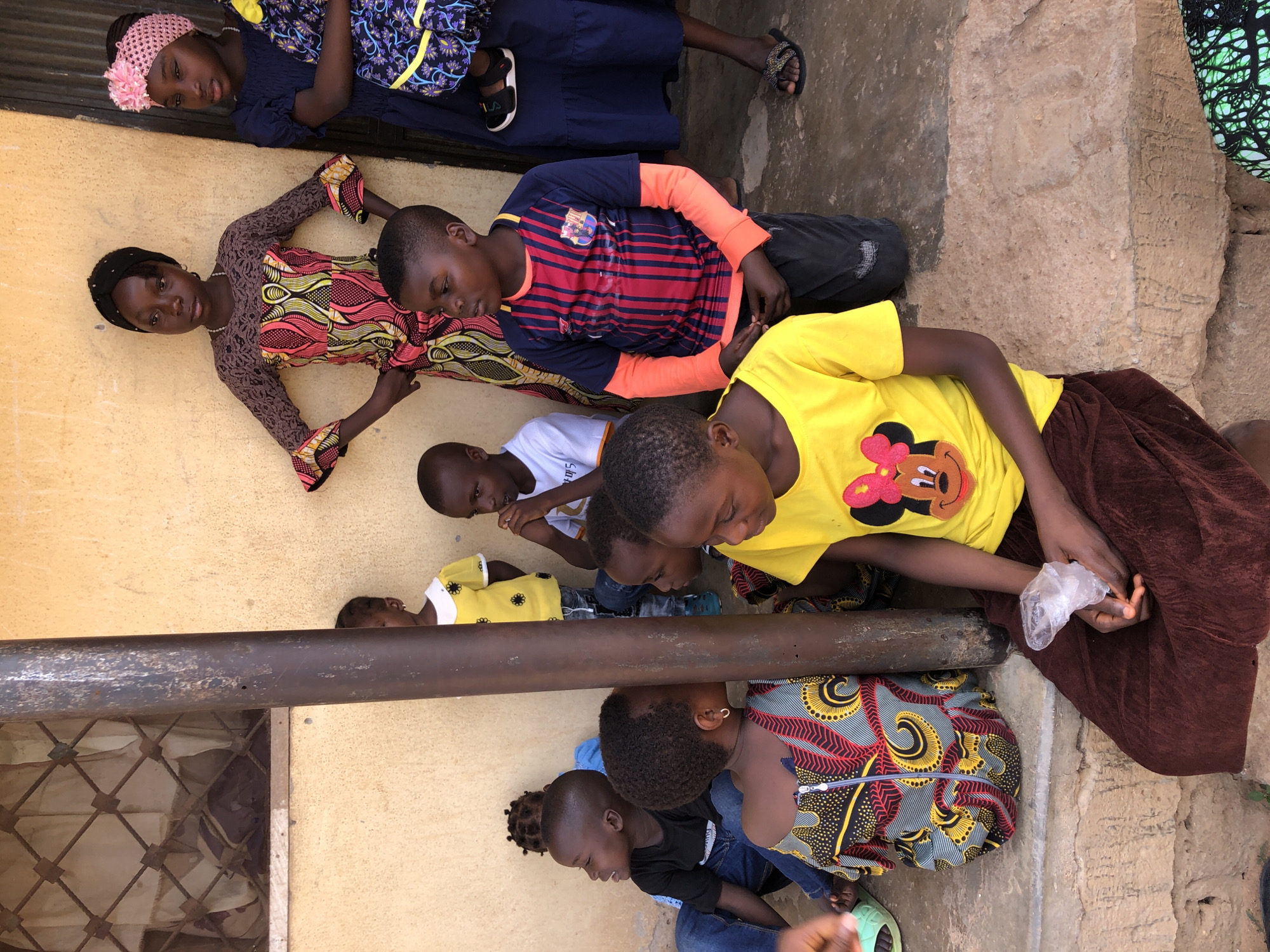 A group of children are gathered on the ground in what appears to be an outdoor area in Akwanga, Nigeria. Several children are lying down, while others are seated or kneeling. They are wearing a variety of casual clothing, including t-shirts, dresses, and jeans. One child is wearing a yellow t-shirt with a cartoon character on it. A building with a metal pipe is visible in the background. The ground appears to be made of concrete or stone. One child is holding a plastic bag in their hands. The setting suggests a yard or common area within a residential or community space.