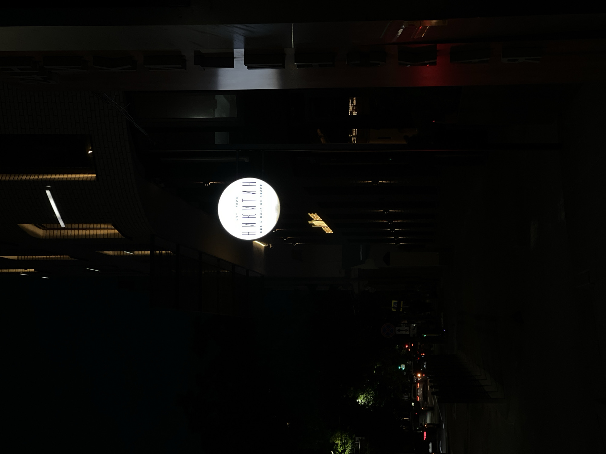 The image shows a nighttime urban scene with a round, illuminated sign that reads "Никитин" in Cyrillic script. The sign also contains smaller text "EST 2024" and "ЕДА И ВИНО ВО ДВОРЕ" (Food and Wine in the Yard). The sign is attached to a building. The environment appears to be a city street at night with faint traffic lights and outlines of buildings visible. The scene is dark, with only a few light sources illuminating the sign and nearby structures. It is not possible to provide information about the city of Svkh Maslovskiy 1 Otdeleniye, Russia, since the only location indicator in the image is on the sign. There are no people visible.