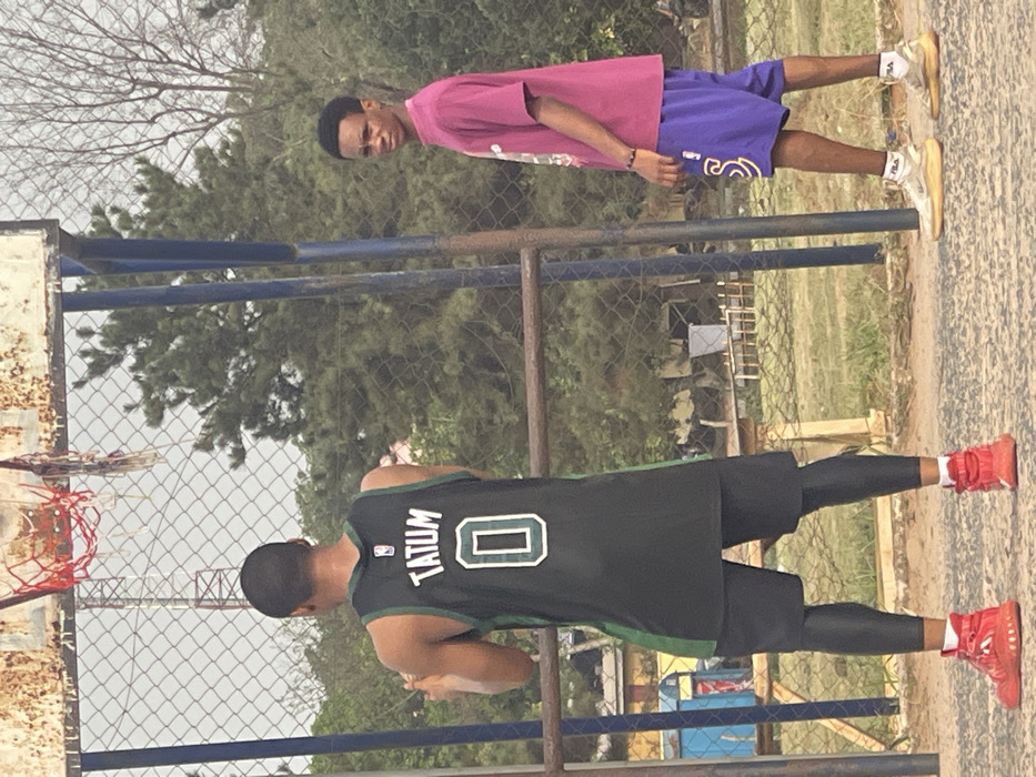 The image depicts an outdoor recreational area in Umunekwu, Nigeria. Two males are present on an unpaved, light brown earth surface. The male on the left is observed from behind, wearing a black and green basketball jersey with "TATUM" and "0" printed on the back, black shorts over black compression leggings, and red high-top athletic shoes. He is positioned facing a basketball hoop. The basketball hoop has a rusted metal backboard frame and a damaged red net, supported by blue poles. The male on the right faces towards the left, looking directly at the camera. He wears a magenta short-sleeved t-shirt, purple basketball shorts with a yellow "S" logo, a dark bracelet on his left wrist, and light-colored sneakers with "FILA" branded socks. A chain-link fence separates the foreground from the background, which features dense green trees and foliage. A distant communication tower is visible through the vegetation on the upper left. No direct interaction between the two individuals is evident.