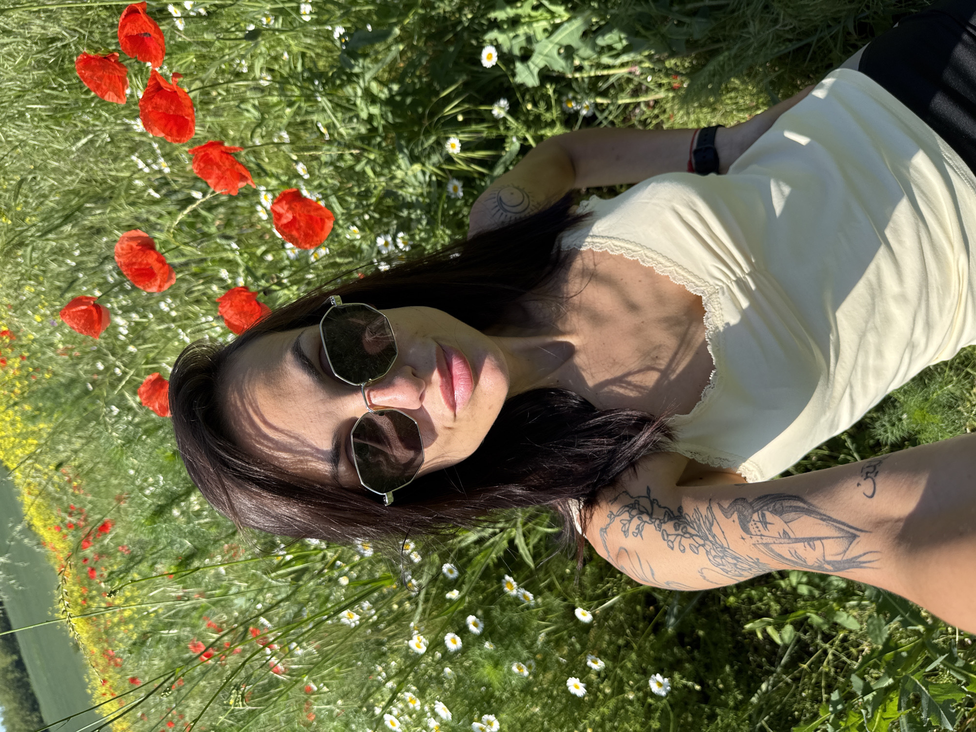 A woman lies in a grassy field interspersed with red poppies and white daisies. She wears sunglasses and a light yellow tank top. Her dark hair is spread around her head. Tattoos are visible on her arm. The photo was likely taken in the Pleven region of Bulgaria, as evidenced by the landscape and flora.