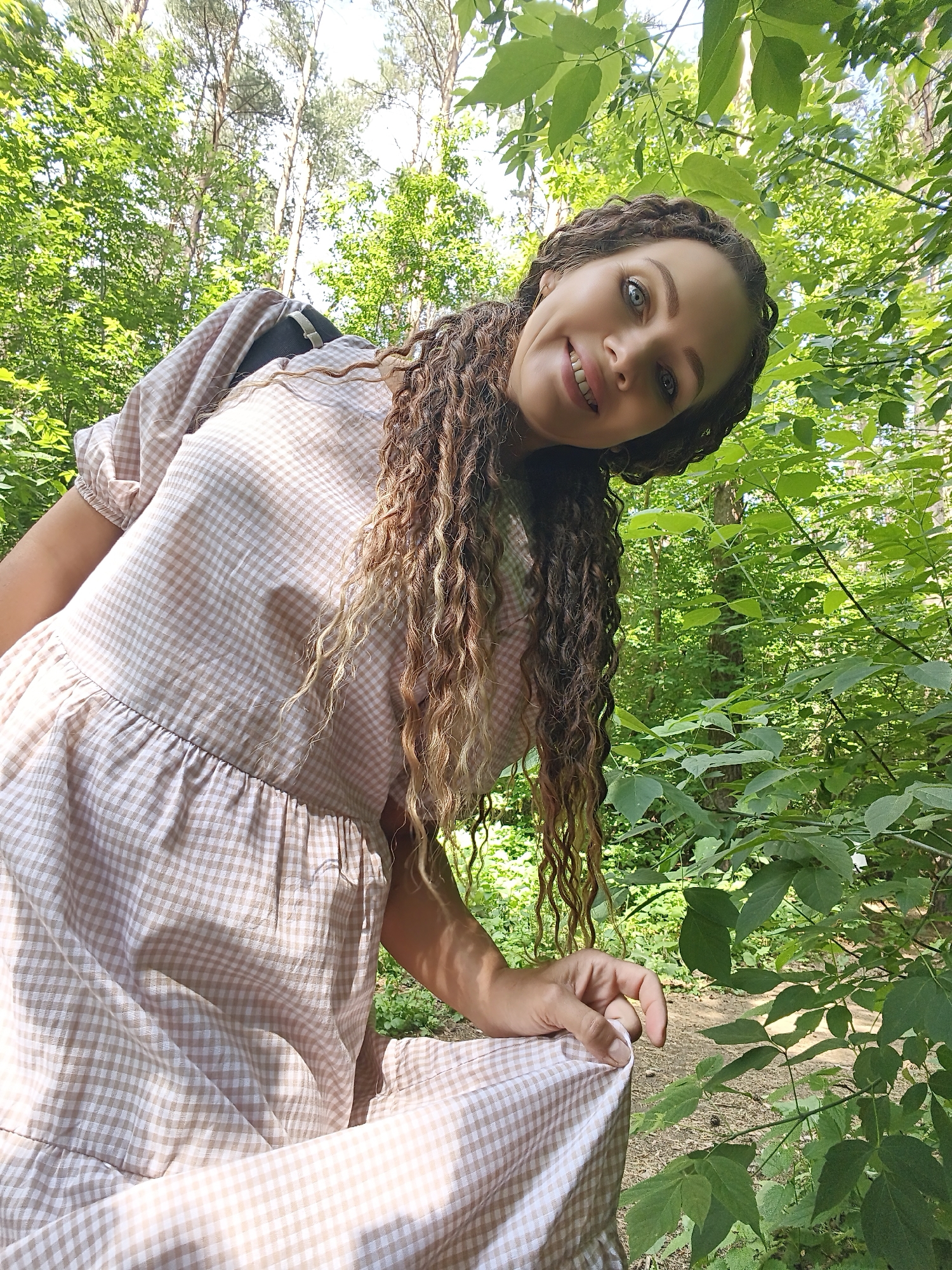 A woman with long, curly brown hair and blue eyes smiles at the camera, leaning down from above. She is wearing a beige and white checkered dress and is outdoors, surrounded by lush green foliage and tall trees. The lighting suggests a sunny day.