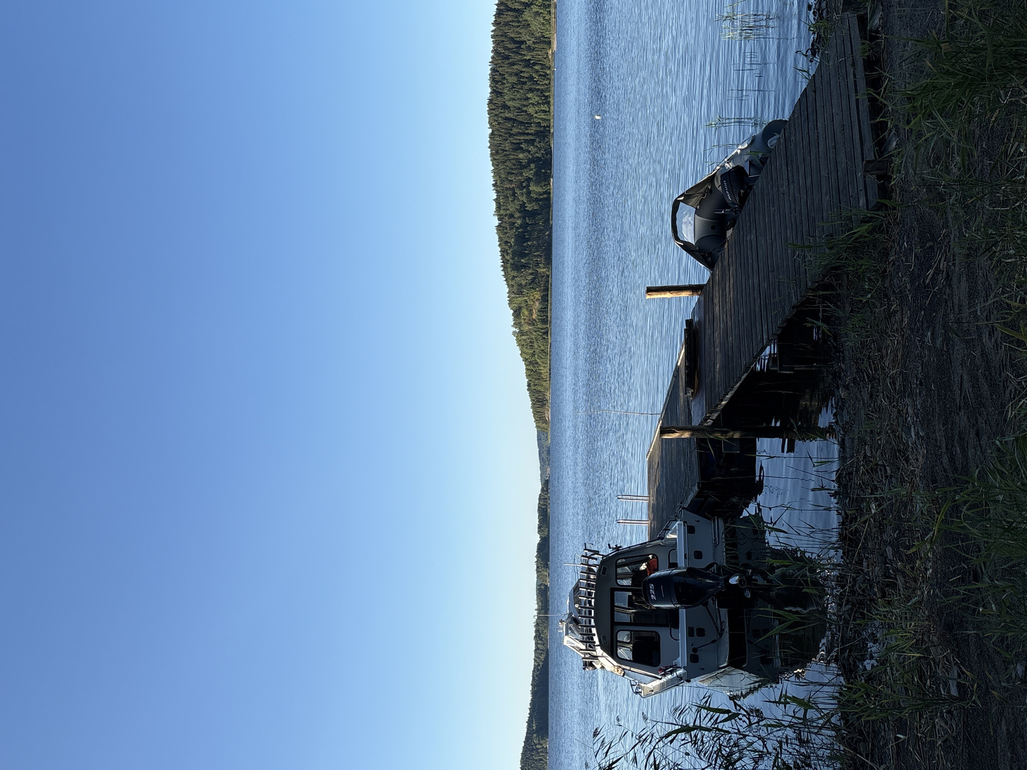 The image depicts a serene lakeside scene in Rautalakhti, Russia. The foreground features a wooden pier extending into a calm body of water. Two boats are docked at the pier: a larger cabin cruiser with a "225" engine visible on the back, and a smaller inflatable boat. The water is blue and slightly rippled, reflecting the clear, bright blue sky. Lush green trees and rolling hills form the landscape on the opposite shore, creating a picturesque backdrop. The overall atmosphere is peaceful and tranquil, suggesting a quiet day by the water.