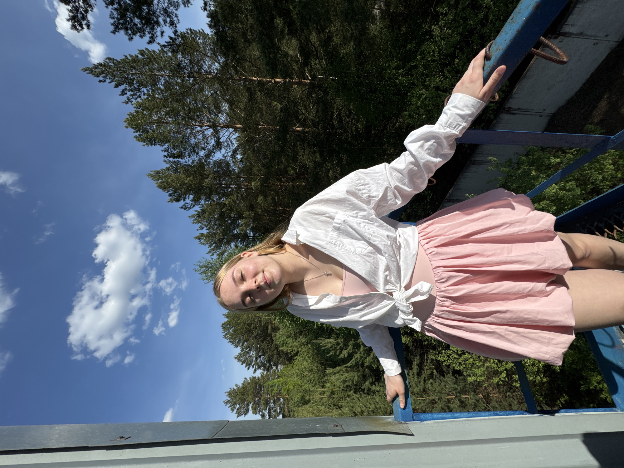 A young woman stands on a blue metal structure outdoors. She is wearing a pink dress with a white shirt tied at the waist. Her left hand holds onto the blue metal, while her right hand rests on top of it. Behind her, a lush forest stretches into the background, and a blue sky with white clouds is visible in the upper left corner. The image appears to be taken from a low angle, tilted slightly. The location is presumed to be Rusalevka, Russia.