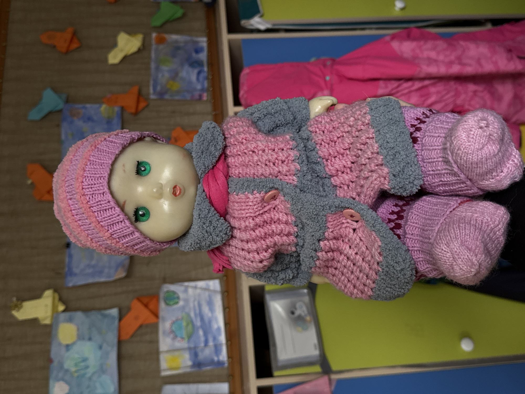 A doll wearing a pink and gray crocheted outfit and hat lies on a surface next to a pink jacket. The doll has green eyes and a pale complexion. The surface appears to be a shelf unit with various colors. Paper crafts in the shapes of airplanes and paintings are scattered on a beige surface to the left. The image does not contain sufficient information to determine its location in Perm, Russia.