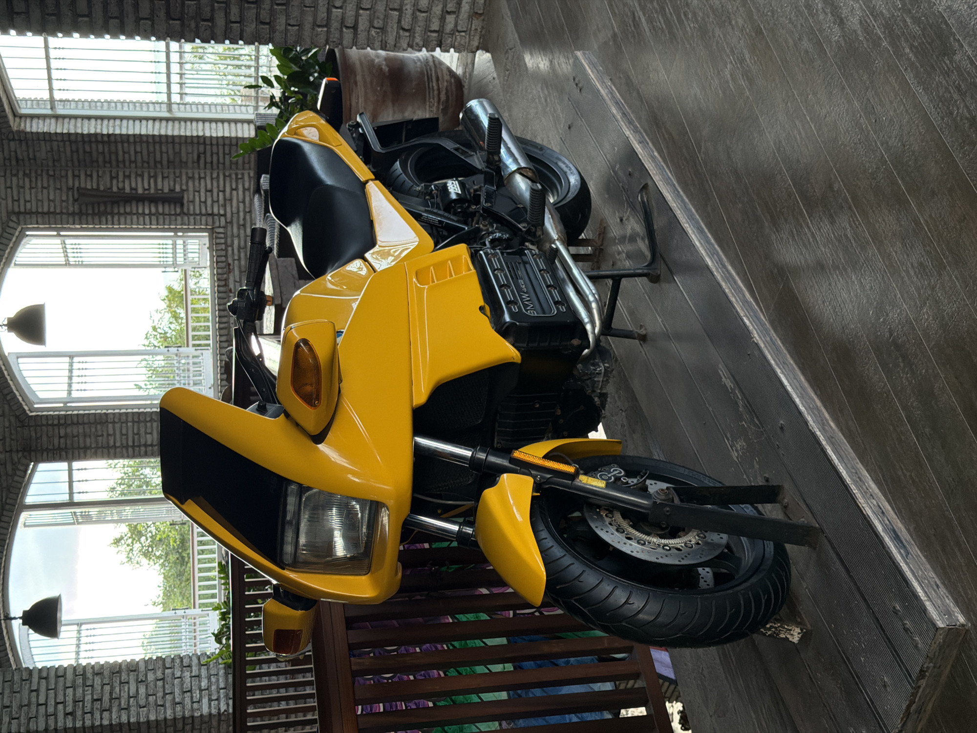 A bright yellow BMW motorcycle is parked on a wooden platform indoors, with large arched windows in the background offering a view of lush green trees and foliage. The motorcycle is the primary subject, facing left and slightly angled towards the viewer. It features a black seat, black handlebars, and a distinctive front fairing. The engine block is visible, with the "BMW" logo clearly marked. The motorcycle is supported by a black kickstand.
The environment appears to be a covered patio or restaurant area with exposed brick walls. The wooden flooring is rough-hewn planks. To the left of the motorcycle, a wooden railing with colorful vertical panels is visible. In the background, through the large windows, a bright, overcast sky is apparent, and the foliage suggests an outdoor natural setting. The scene is static and there are no people or animals present. The lighting is even, suggesting it's daytime with diffused natural light.