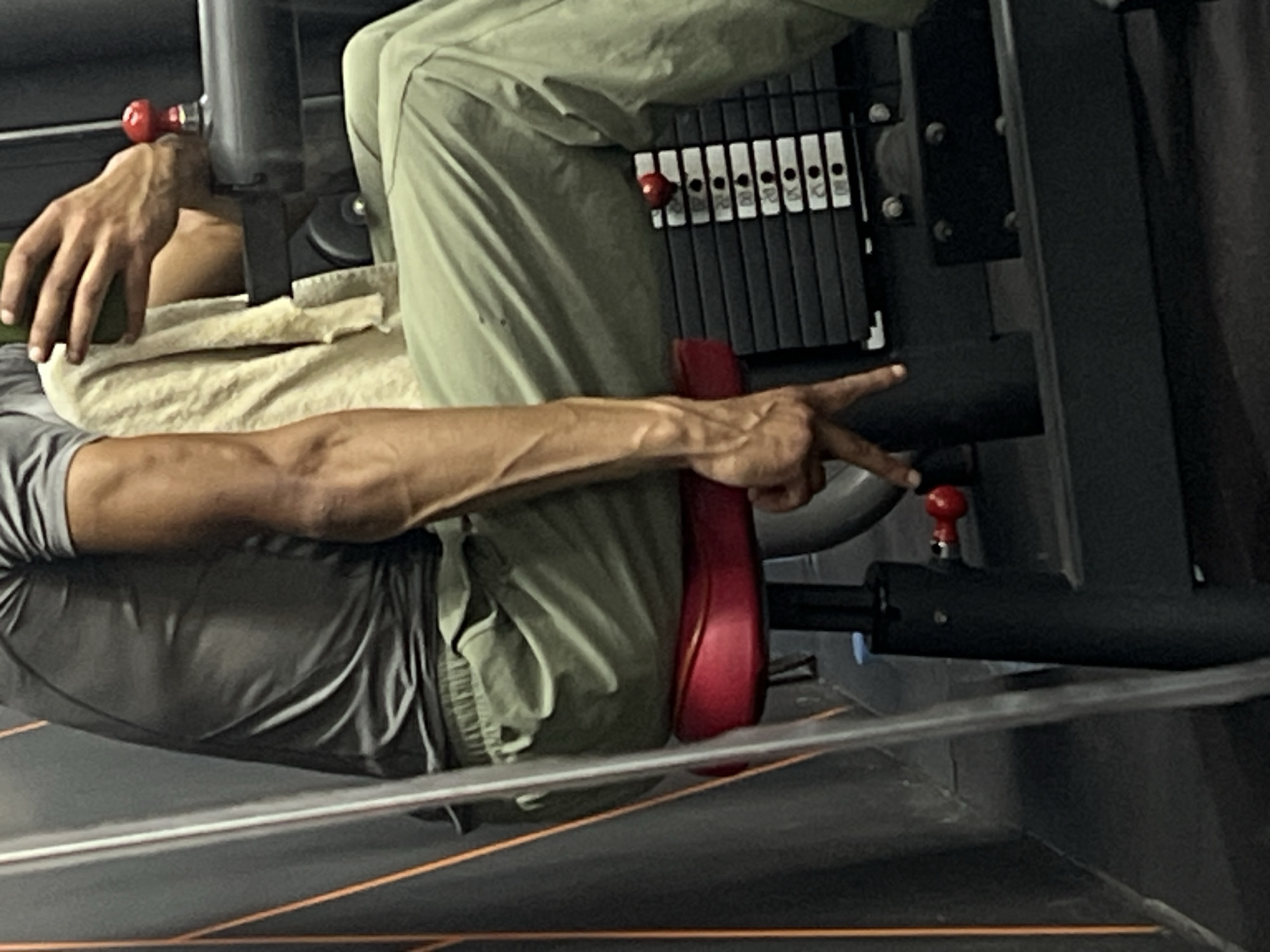 This image captures a person engaged in a leg exercise at a gym, likely in M'Diq, Morocco. The photo is oriented ninety degrees counter-clockwise from the upright position, showing the individual lying on their back on a gym machine, possibly a leg press.
The subject is partially visible, wearing grey shorts and light green long pants or track bottoms. A grey t-shirt is also visible on their upper body. A beige towel is draped beneath their torso. Their left hand rests casually, while their right arm is extended towards the machine's weight stack. Prominent veins are visible on their right forearm, indicating muscle definition. With their right hand, they are making a two-finger gesture, possibly pointing at the weight selected on the stack or interacting with a mechanism.
The gym machine is robust, primarily black and grey, featuring a vertical weight stack composed of multiple black plates. Numbers indicating weight increments are visible on the plates, including "20", "25", "30", "35", "40", "45", "50", and "55", likely in kilograms. A red pin is inserted into one of these plates to select the desired weight. There are also distinct red footrests or pads where the person's feet would push. Another small red knob or lever is visible near the top of the machine frame, and another red adjustment pin is lower down.
The setting is an indoor gym, characterized by dark equipment and a dark floor with orange lines, suggesting a dedicated workout space. The lighting is artificial, typical of an indoor environment. The person appears focused on their exercise, with no specific emotional expression discernible.