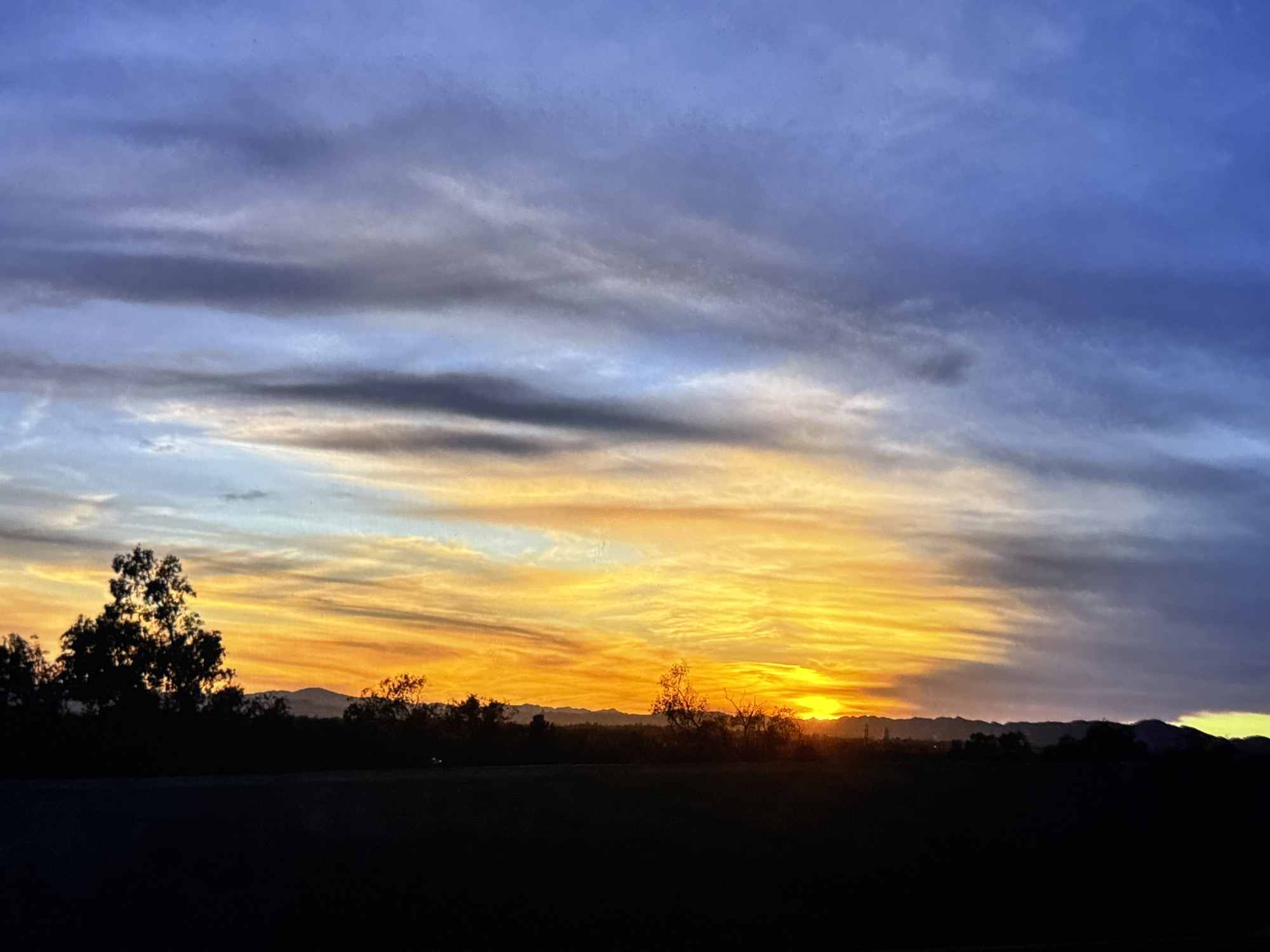 This is a photograph of a sunset over a silhouetted landscape in Los Angeles, California, United States of America. The sky displays a gradient of colors, transitioning from a deep blue at the top to vibrant oranges and yellows near the horizon. Wispy clouds are visible throughout the sky, catching the light of the setting sun. In the foreground, a dark silhouette of trees and distant mountains stretches across the frame. The lower portion of the image is a solid black silhouette, suggesting a foreground element like a road or flat terrain. The sun is visible as a bright orb on the horizon, partially obscured by the silhouetted landscape.