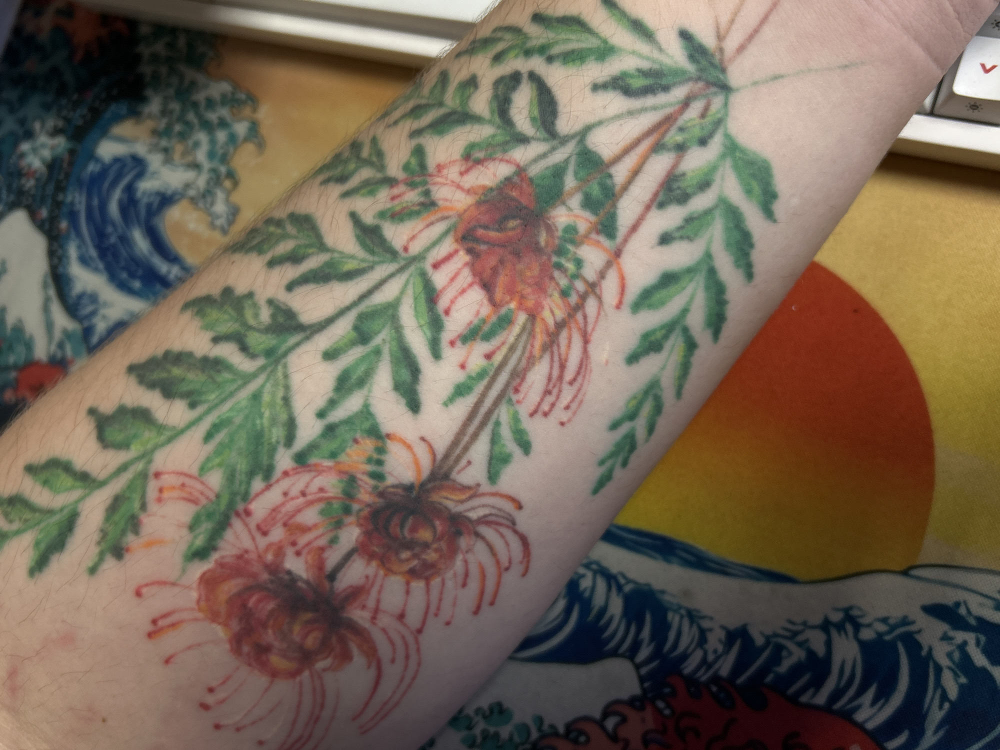 A close-up photograph features a person's forearm adorned with a tattoo of red spider lilies and green foliage. The forearm is pale and slightly hairy. The tattoo is detailed, depicting three red spider lilies with their characteristic spidery petals and slender stems, surrounded by stylized green leaves. The background includes a portion of a desk mat with a print of a stylized wave pattern in blue and white, and part of a keyboard. The location is not discernible from the image.