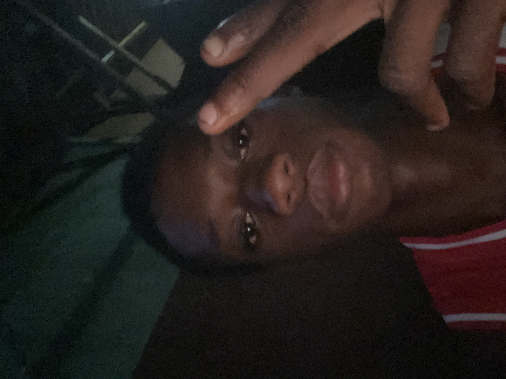 A close-up, dimly lit photograph shows a person with dark skin making a hand gesture near their face. The person is wearing a red and white striped garment. The background is dark and out of focus, with a vague suggestion of some objects. The location of Nkpor, Nigeria, cannot be verified from the image.